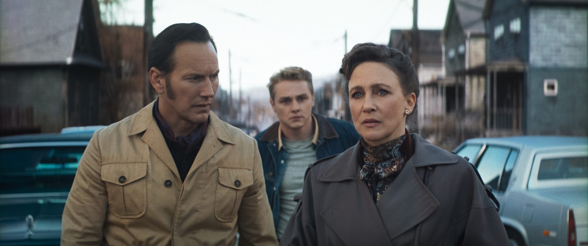 a trio of people walking together on a street lined with old houses and parked cars the man on the left wears a beige jacket the young man in the middle has lightcolored hair and denim while the woman on the right is dressed in a dark coat and scarf all displaying serious expressions digital production A trio of people walking together on a street lined with old houses and parked cars. The man on the left wears a beige jacket, the young man in the middle has light-colored hair and denim, while the woman on the right is dressed in a dark coat and scarf, all displaying serious expressions.