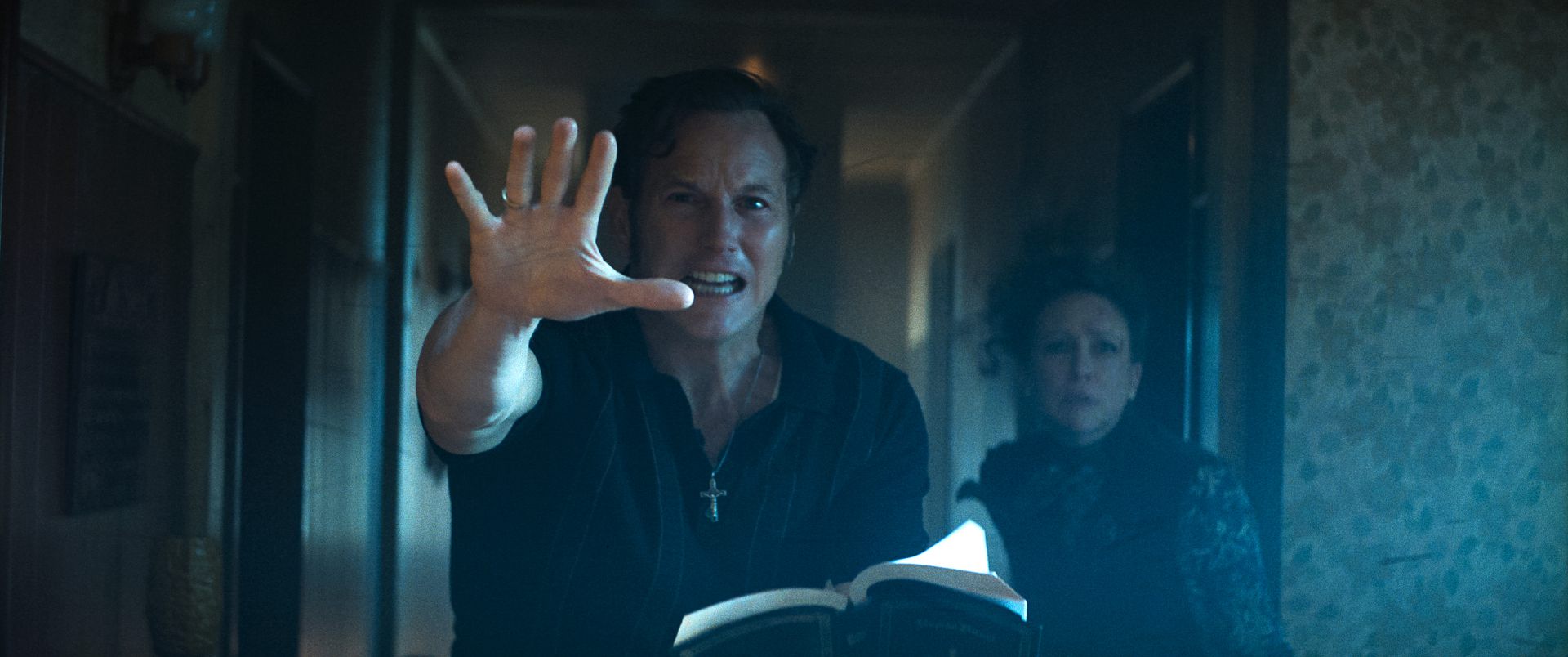 a man in a dark shirt and cross necklace extends his hand while holding an open book showing urgency a woman stands behind him with a concerned expression both in a dimly lit corridor with floral wallpaper digital production A man in a dark shirt and cross necklace extends his hand while holding an open book, showing urgency. A woman stands behind him with a concerned expression, both in a dimly lit corridor with floral wallpaper.
