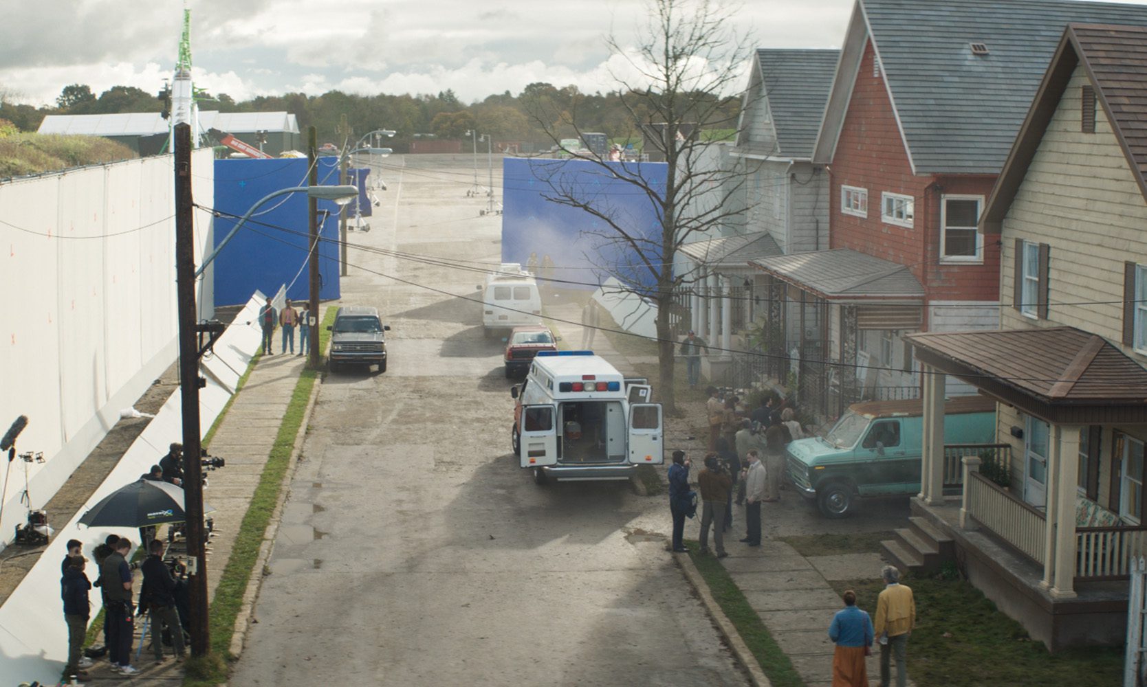 a street scene depicting a film set with blue backdrops and crew members nearby an ambulance is parked with its doors open while a vintage car is positioned beside a house the setting is overcast suggesting an industrial area digital production A street scene depicting a film set, with blue backdrops and crew members nearby. An ambulance is parked with its doors open, while a vintage car is positioned beside a house. The setting is overcast, suggesting an industrial area.