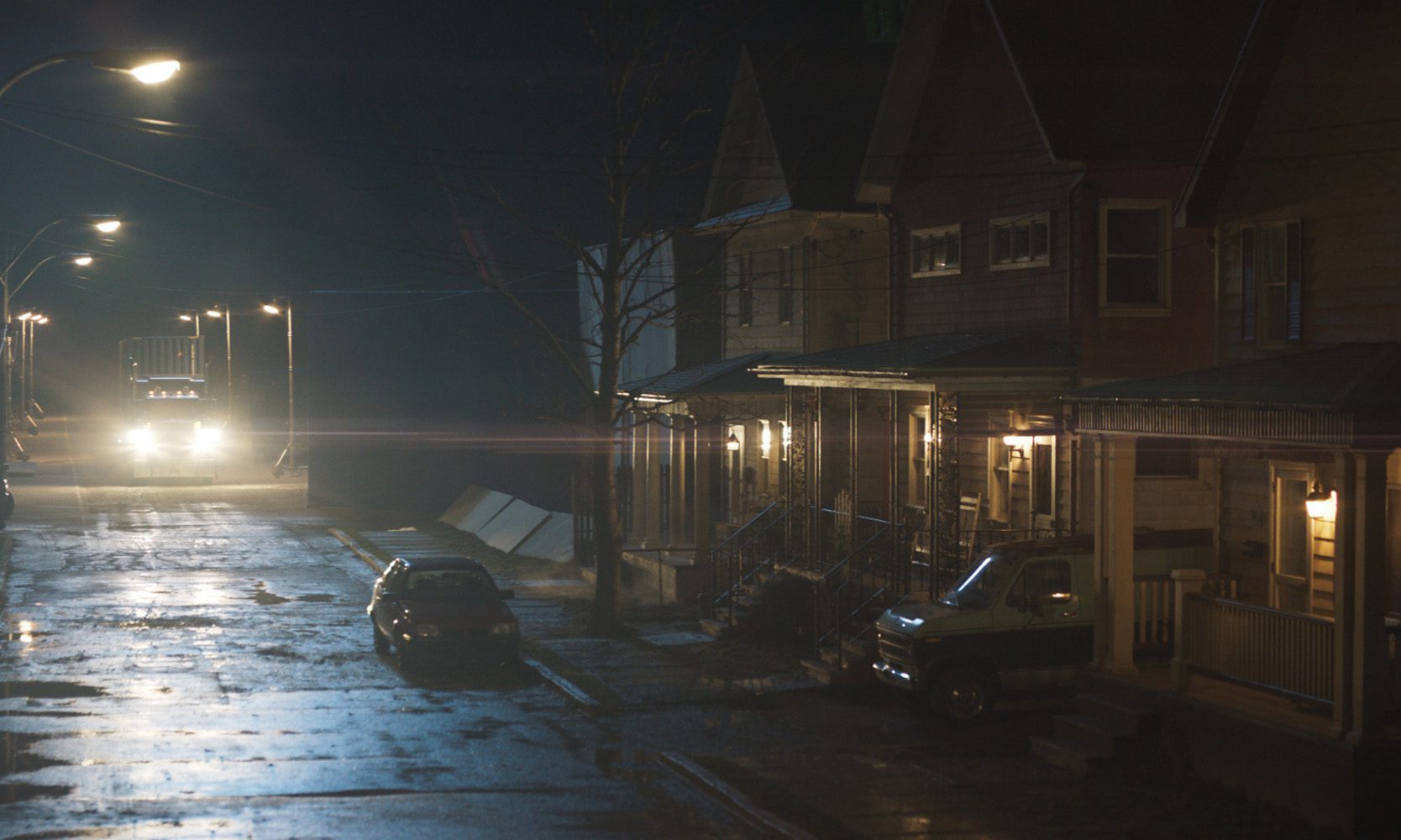 a dimly lit street at night with vintage houses lining both sides a large trucks headlights illuminate the wet pavement reflecting a yellow glow a parked car is visible on the left with shadows emphasizing the quiet rural atmosphere digital production A dimly lit street at night with vintage houses lining both sides. A large truck's headlights illuminate the wet pavement, reflecting a yellow glow. A parked car is visible on the left, with shadows emphasizing the quiet, rural atmosphere.
