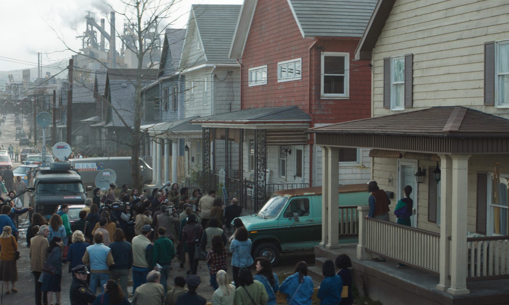 a crowd of people congregates on a street in front of houses with news vans parked nearby some individuals are visible on porches while others stand amidst the gathering set against an industrial backdrop digital production A crowd of people congregates on a street in front of houses, with news vans parked nearby. Some individuals are visible on porches, while others stand amidst the gathering, set against an industrial backdrop.