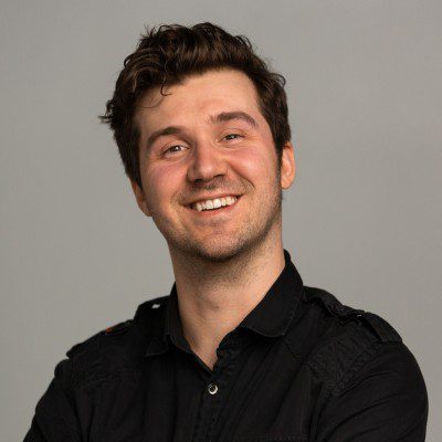 a smiling young man with short wavy hair wearing a black buttonup shirt standing against a neutral gray background he is looking directly at the camera with a friendly expression arms crossed digital production A smiling young man with short, wavy hair, wearing a black button-up shirt, standing against a neutral gray background. He is looking directly at the camera with a friendly expression, arms crossed.