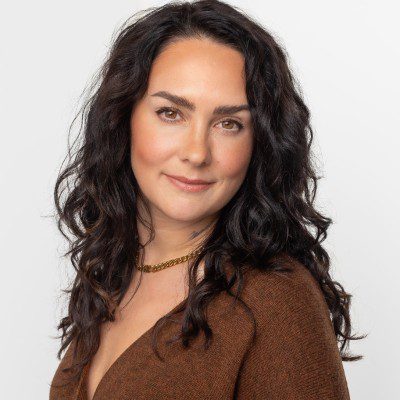 a woman with long curly dark hair wearing a brown sweater and a gold necklace posing against a plain white background she has a subtle smile and a relaxed expression creating a warm and approachable appearance digital production A woman with long, curly dark hair wearing a brown sweater and a gold necklace, posing against a plain white background. She has a subtle smile and a relaxed expression, creating a warm and approachable appearance.