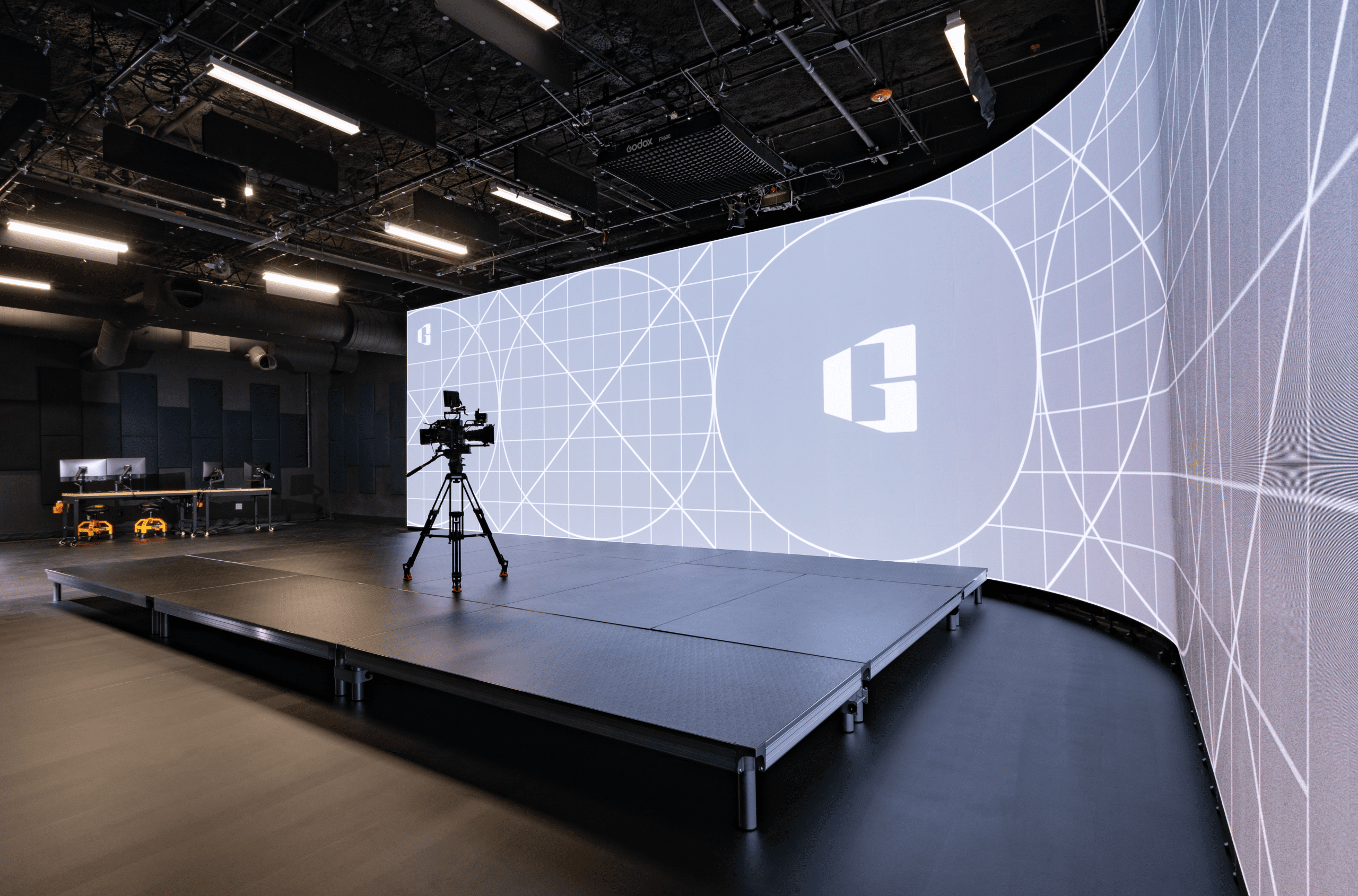 a modern studio setup featuring a large curved led screen displaying a grid pattern in the foreground a professional camera on a tripod is positioned on a raised platform with a sleek workspace visible in the background digital production A modern studio setup featuring a large curved LED screen displaying a grid pattern. In the foreground, a professional camera on a tripod is positioned on a raised platform, with a sleek workspace visible in the background.
