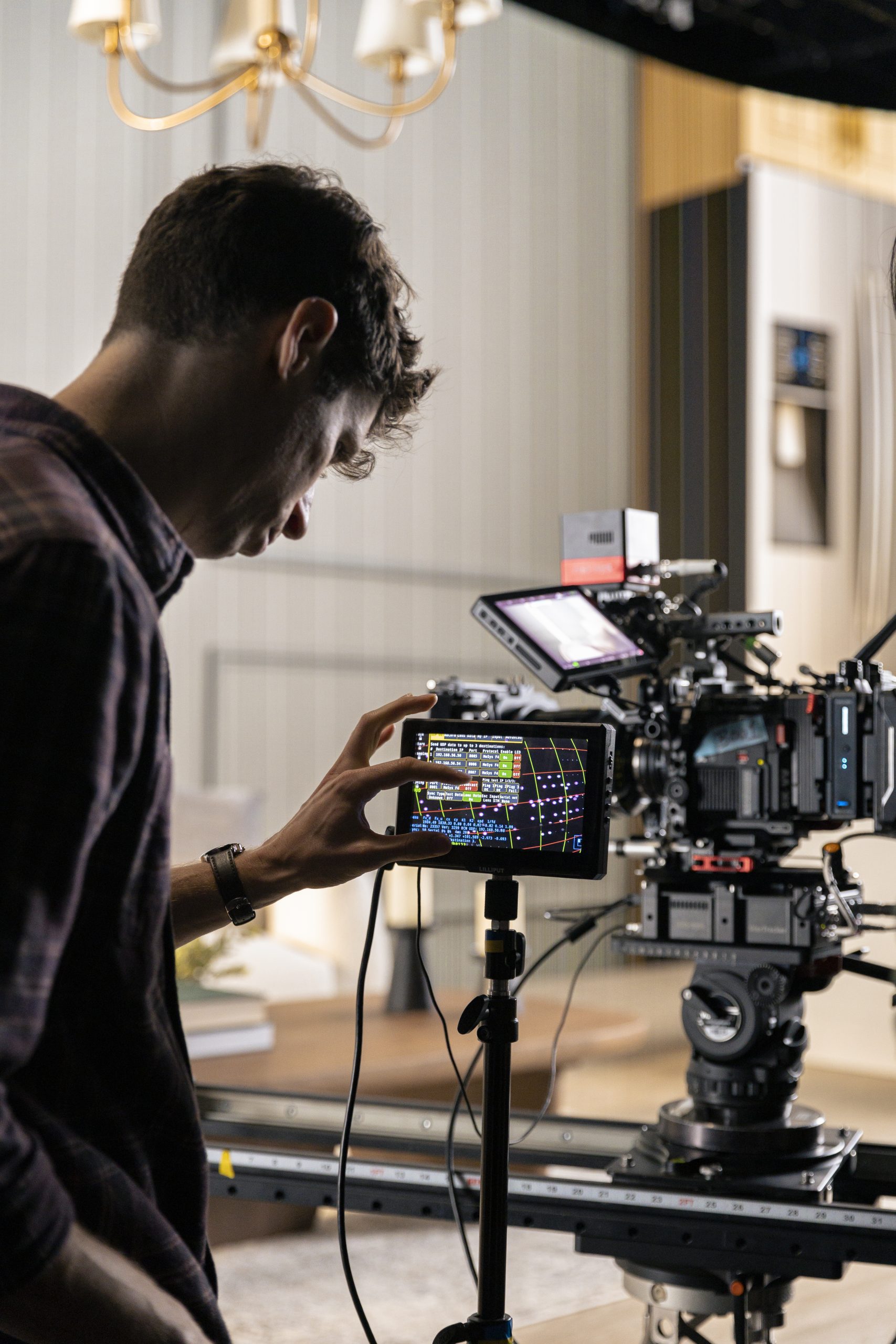 a film crew member adjusting camera settings on a digital monitor attached to a camera on a tripod with a softly lit interior background suggesting a production set digital production A film crew member adjusting camera settings on a digital monitor attached to a camera on a tripod, with a softly lit interior background suggesting a production set.