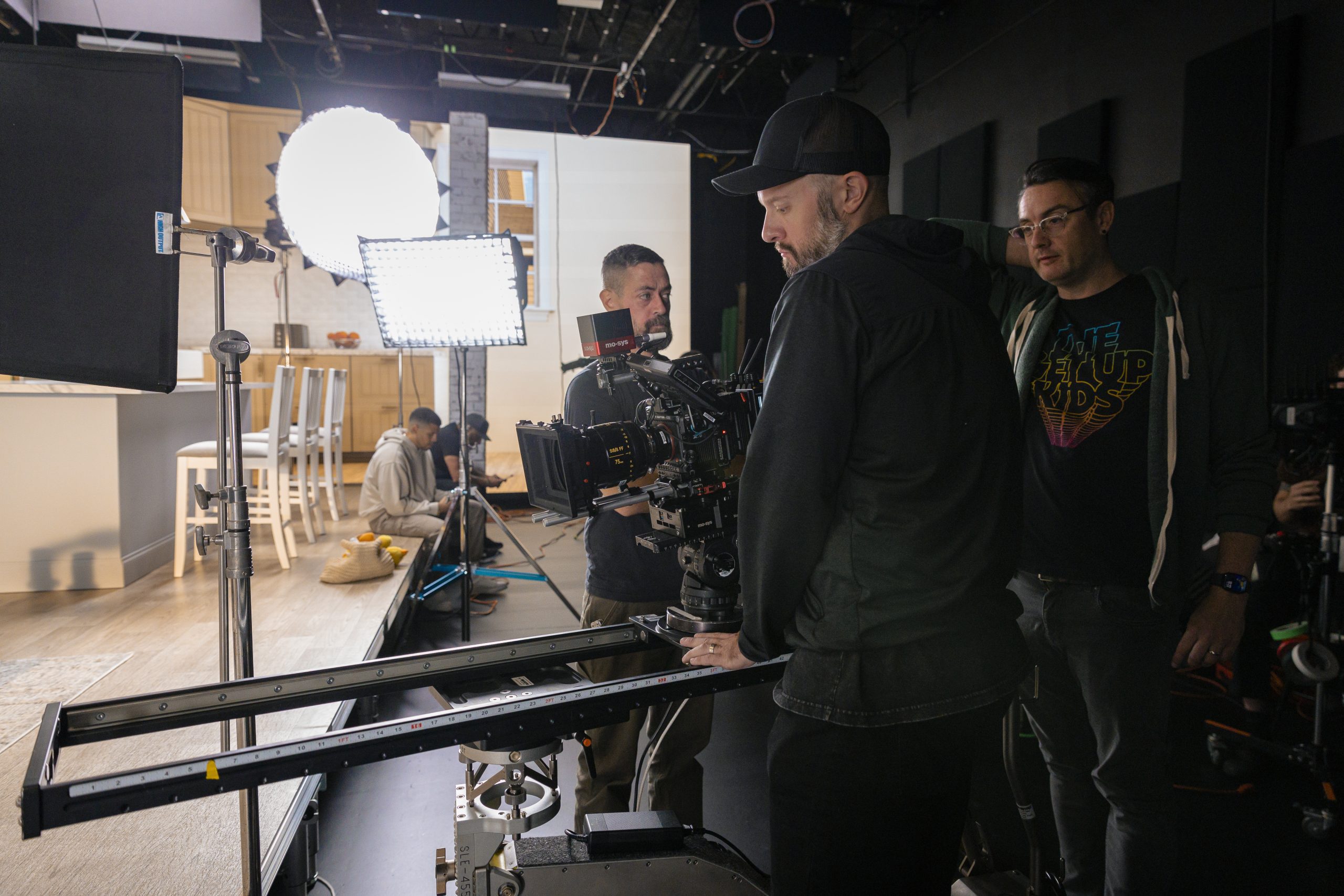 a film crew setting up equipment in a studio one person adjusts a camera on a sliding rig while others assist with lighting and monitor screens a kitchen set is visible in the background creating a professional filming environment digital production A film crew setting up equipment in a studio. One person adjusts a camera on a sliding rig while others assist with lighting and monitor screens. A kitchen set is visible in the background, creating a professional filming environment.
