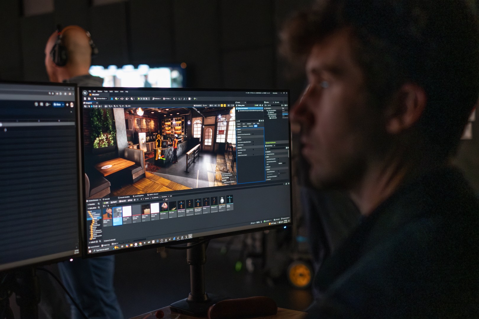 a focused individual sitting at a desk observing a computer screen displaying a detailed rendering of an interior space while a person with headphones is visible in the background digital production A focused individual sitting at a desk, observing a computer screen displaying a detailed rendering of an interior space, while a person with headphones is visible in the background.