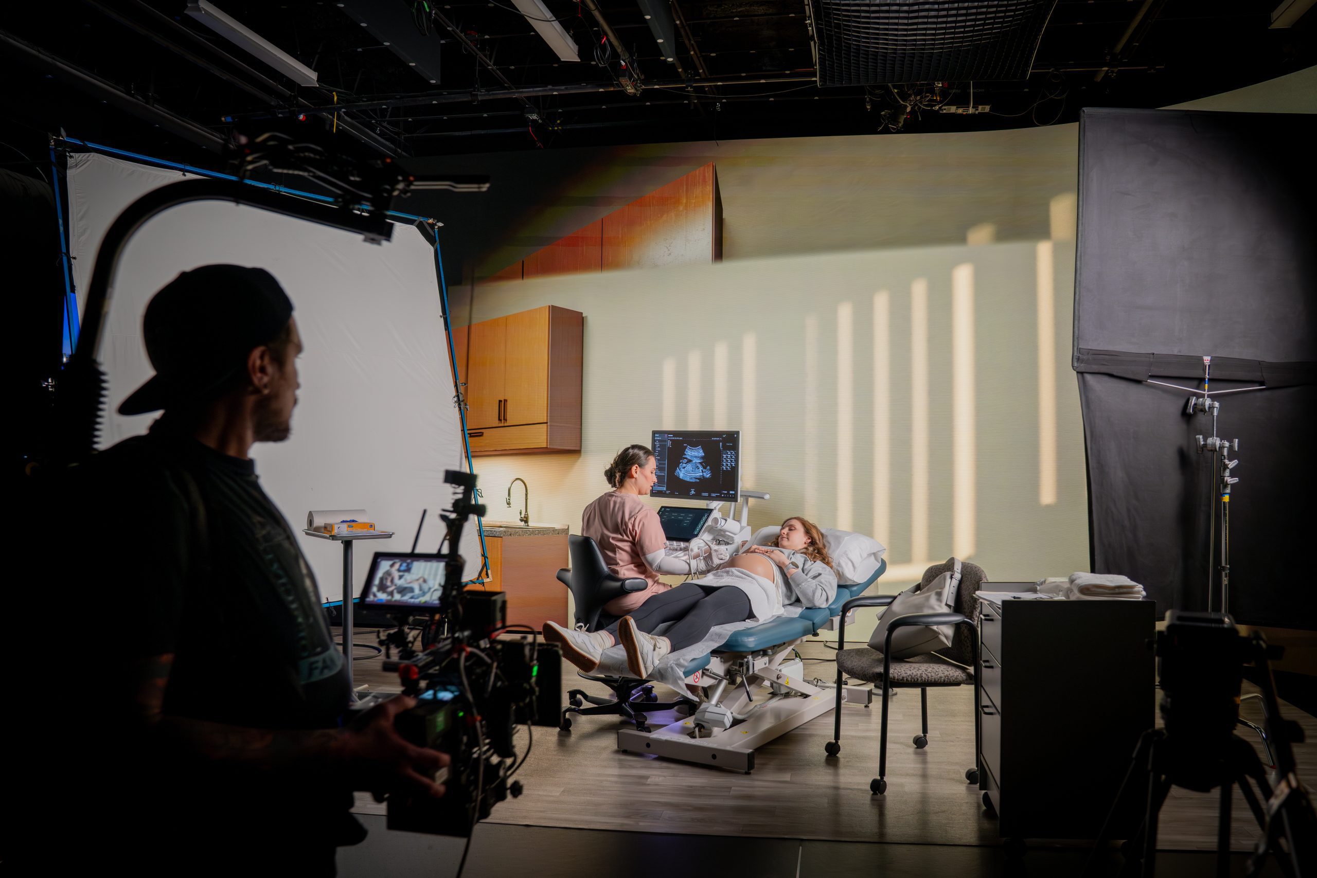 a film crew capturing a scene in a hospital set featuring an actor lying on a hospital bed while another actor interacts with a medical monitor the set has modern elements and soft lighting with a crew member visible in the foreground focusing on the shoot digital production A film crew capturing a scene in a hospital set, featuring an actor lying on a hospital bed while another actor interacts with a medical monitor. The set has modern elements and soft lighting, with a crew member visible in the foreground focusing on the shoot.
