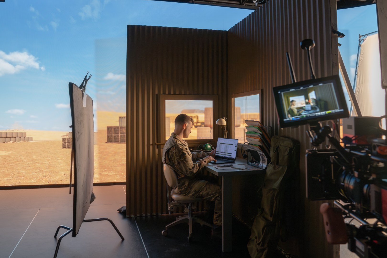 a soldier in military fatigues sitting at a desk working on a laptop the background features a desert landscape with cargo containers visible a camera and equipment are set up nearby indicating a filming or recording session digital production A soldier in military fatigues sitting at a desk, working on a laptop. The background features a desert landscape with cargo containers visible. A camera and equipment are set up nearby, indicating a filming or recording session.