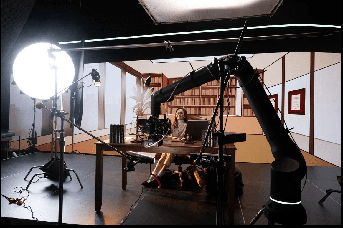 a woman sitting at a desk in a studio with bookshelves in the background the scene is illuminated by a large softbox light and a robotic camera arm studio equipment is arranged around her creating a professional filming setup digital production A woman sitting at a desk in a studio with bookshelves in the background. The scene is illuminated by a large softbox light and a robotic camera arm. Studio equipment is arranged around her, creating a professional filming setup.