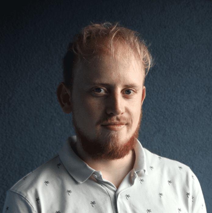 a closeup portrait of a young man with light brown hair and a beard wearing a white shirt with small dark patterns he poses against a textured blue wall giving a subtle smile digital production A close-up portrait of a young man with light brown hair and a beard, wearing a white shirt with small dark patterns. He poses against a textured blue wall, giving a subtle smile.