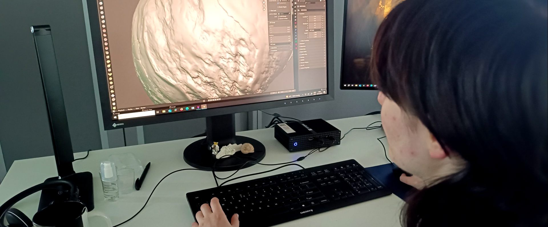 a person seated at a desk focused on a computer screen displaying intricate 3d modeling software various tools and a glass of water are placed on the desk beside a keyboard digital production A person seated at a desk, focused on a computer screen displaying intricate 3D modeling software. Various tools and a glass of water are placed on the desk beside a keyboard.