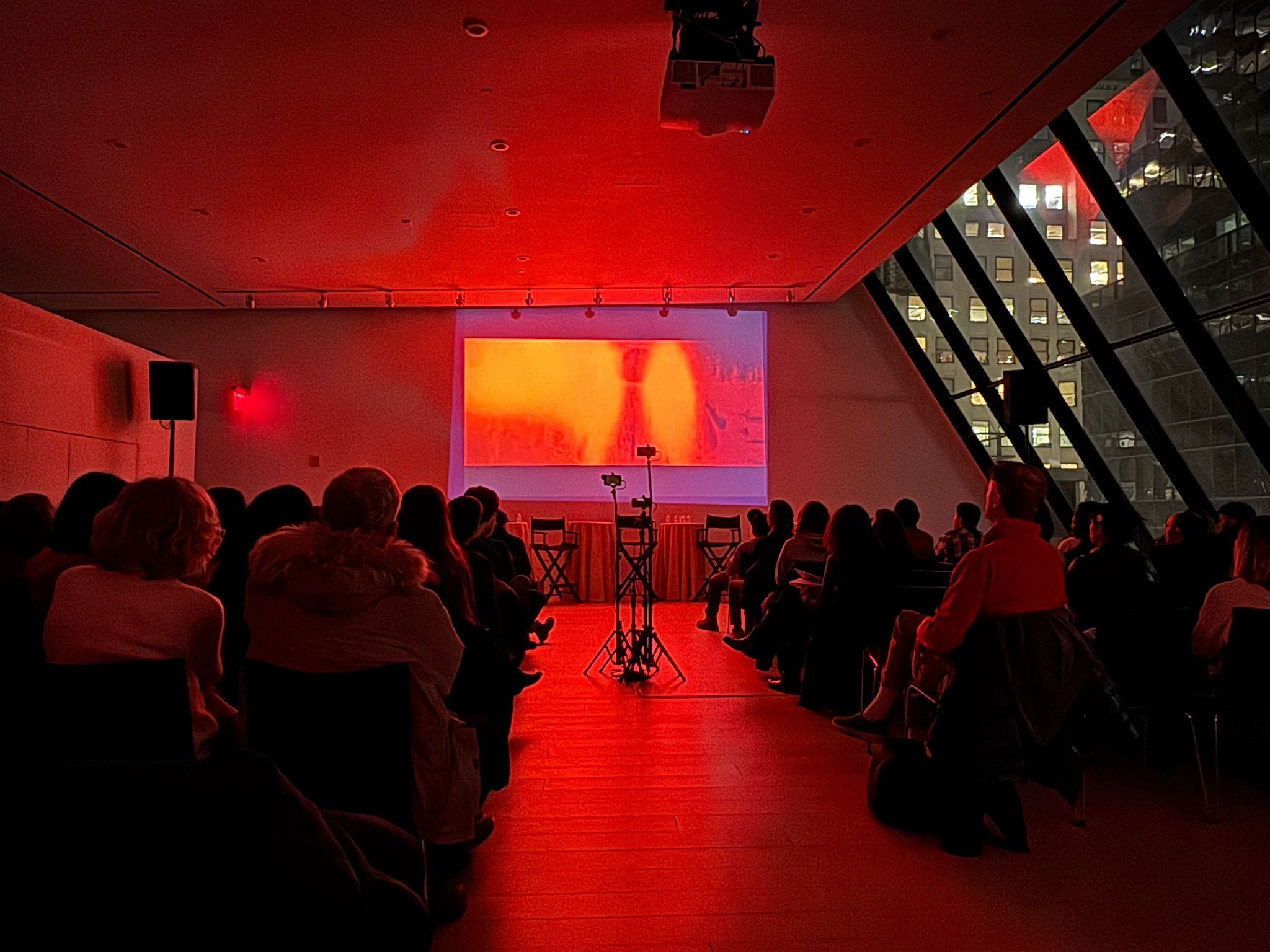 a dimly lit room with red lighting where an audience is seated facing a screen displaying a vibrant visual large windows show the city skyline at night digital production A dimly lit room with red lighting where an audience is seated, facing a screen displaying a vibrant visual. Large windows show the city skyline at night.
