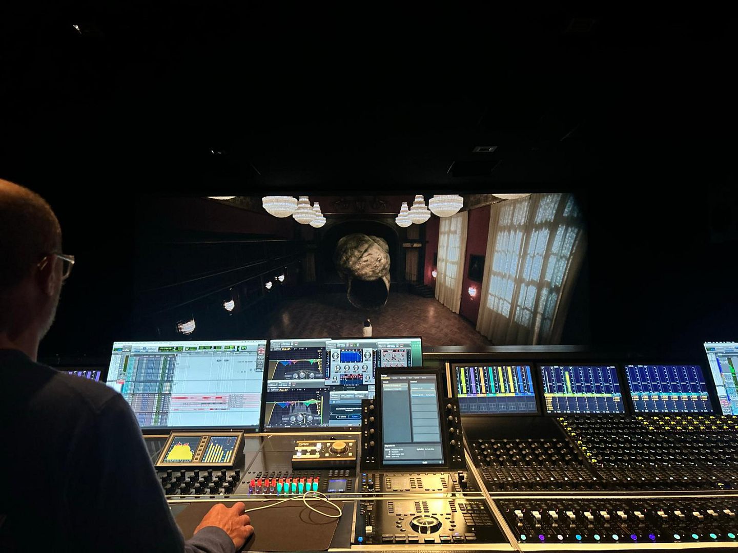 a sound engineer adjusting audio settings at a mixing console in a dark studio in the background a large screen displays an atmospheric scene of a dimly lit hallway with ornate chandeliers and a large spherical object digital production A sound engineer adjusting audio settings at a mixing console in a dark studio. In the background, a large screen displays an atmospheric scene of a dimly lit hallway with ornate chandeliers and a large spherical object.