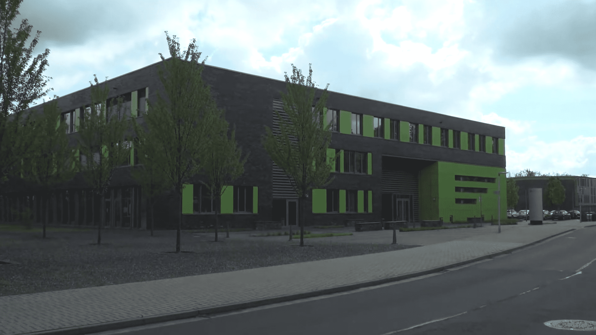 a modern building featuring a dark facade with large green windows surrounded by small trees the structure has a minimalist design and is located beside a paved road with parking spaces visible in the background digital production A modern building featuring a dark facade with large green windows, surrounded by small trees. The structure has a minimalist design and is located beside a paved road with parking spaces visible in the background.