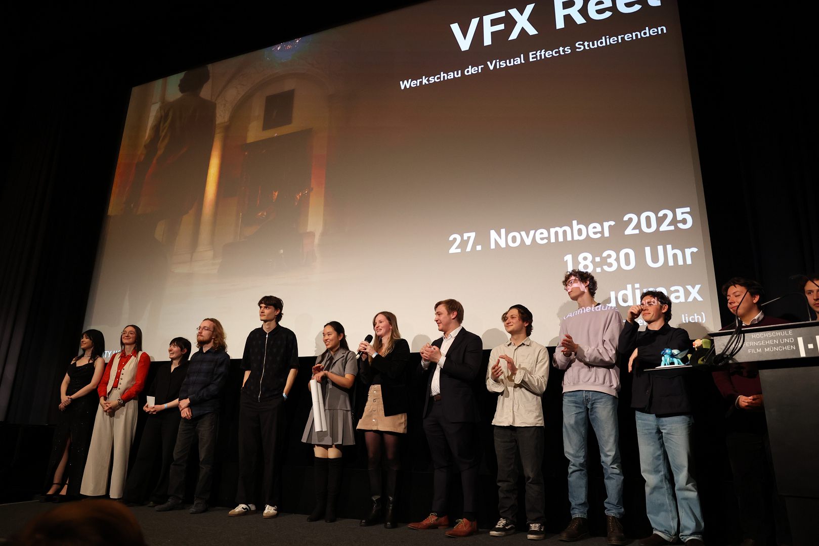 a group of students stands on stage during a visual effects showcase event they are smiling and applauding in front of a large screen displaying event details the background has a dark setting with bright lighting directed at the stage digital production A group of students stands on stage during a visual effects showcase event. They are smiling and applauding in front of a large screen displaying event details. The background has a dark setting with bright lighting directed at the stage.