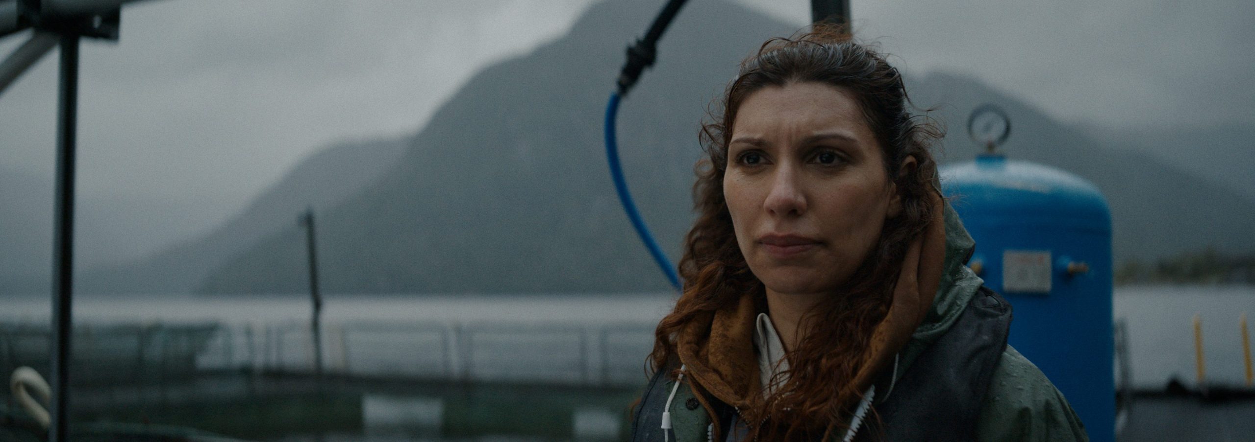 a woman with wavy brown hair and serious expression stands near water dressed in a warm jacket in the background misty mountains and a blue tank are visible under cloudy skies digital production A woman with wavy brown hair and serious expression stands near water, dressed in a warm jacket. In the background, misty mountains and a blue tank are visible under cloudy skies.