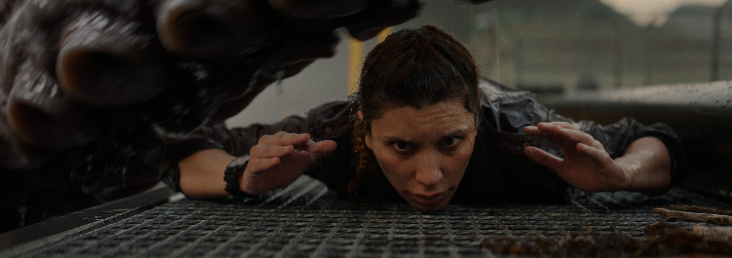 a woman crouching low on a metal grate looking determined and alert as a large creature looms above her partially obscured the setting appears dimly lit intensifying the sense of danger in the scene digital production A woman crouching low on a metal grate, looking determined and alert, as a large creature looms above her, partially obscured. The setting appears dimly lit, intensifying the sense of danger in the scene.