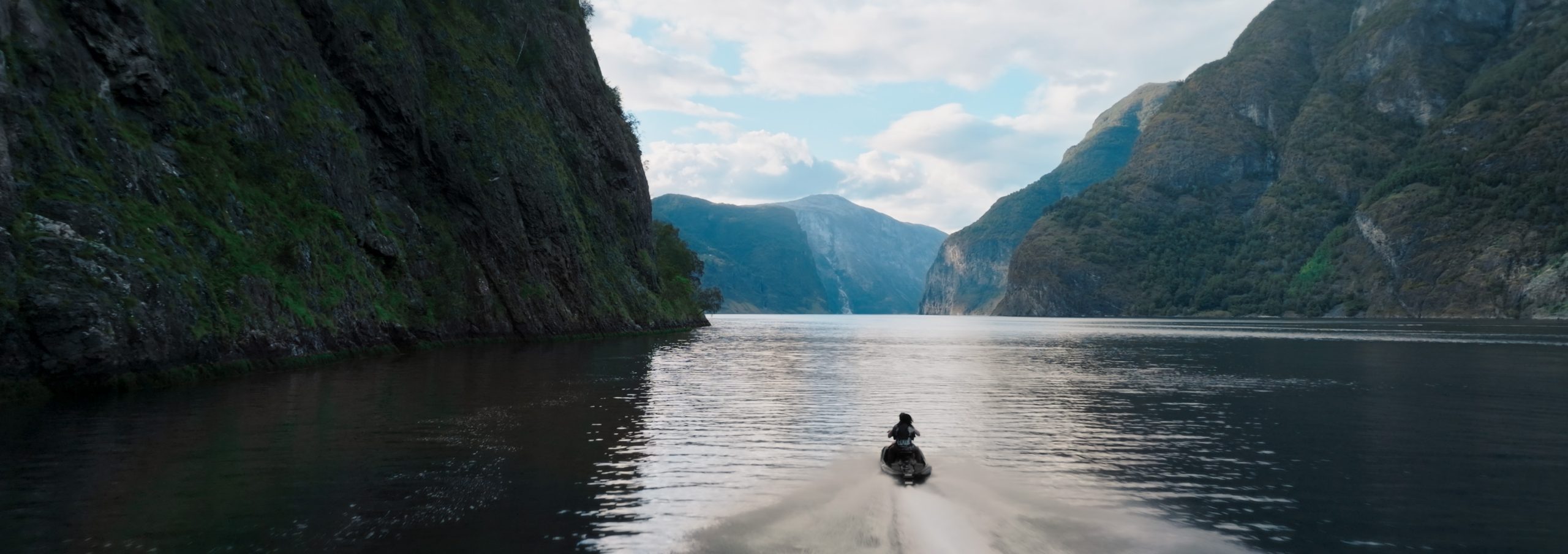 a person riding a jet ski through a tranquil fjord surrounded by steep green cliffs and mountains under a cloudy sky with smooth water reflecting the landscape digital production A person riding a jet ski through a tranquil fjord, surrounded by steep green cliffs and mountains under a cloudy sky, with smooth water reflecting the landscape.