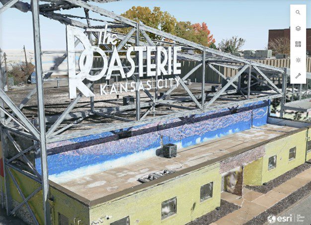 an aerial view of a coffee roasting facility called the roasterie featuring a colorful mural on the buildings wall the structure shows a partially open roof with a metal framework surrounded by greenery digital production An aerial view of a coffee roasting facility called 'The Roasterie', featuring a colorful mural on the building's wall. The structure shows a partially open roof with a metal framework, surrounded by greenery.
