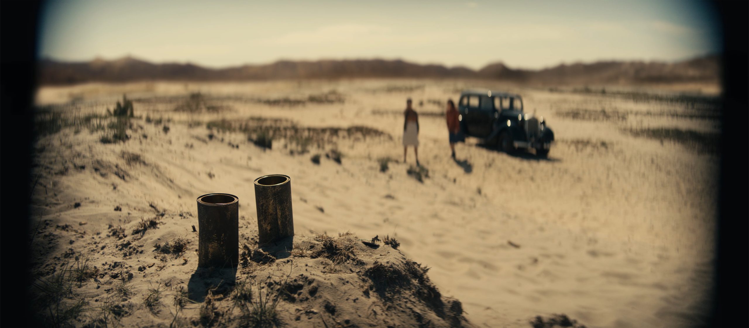 two cylindrical objects on a sandy terrain with a vintage car parked in the background two figures stand near the vehicle against a backdrop of distant hills and an expansive blue sky digital production Two cylindrical objects on a sandy terrain, with a vintage car parked in the background. Two figures stand near the vehicle against a backdrop of distant hills and an expansive blue sky.