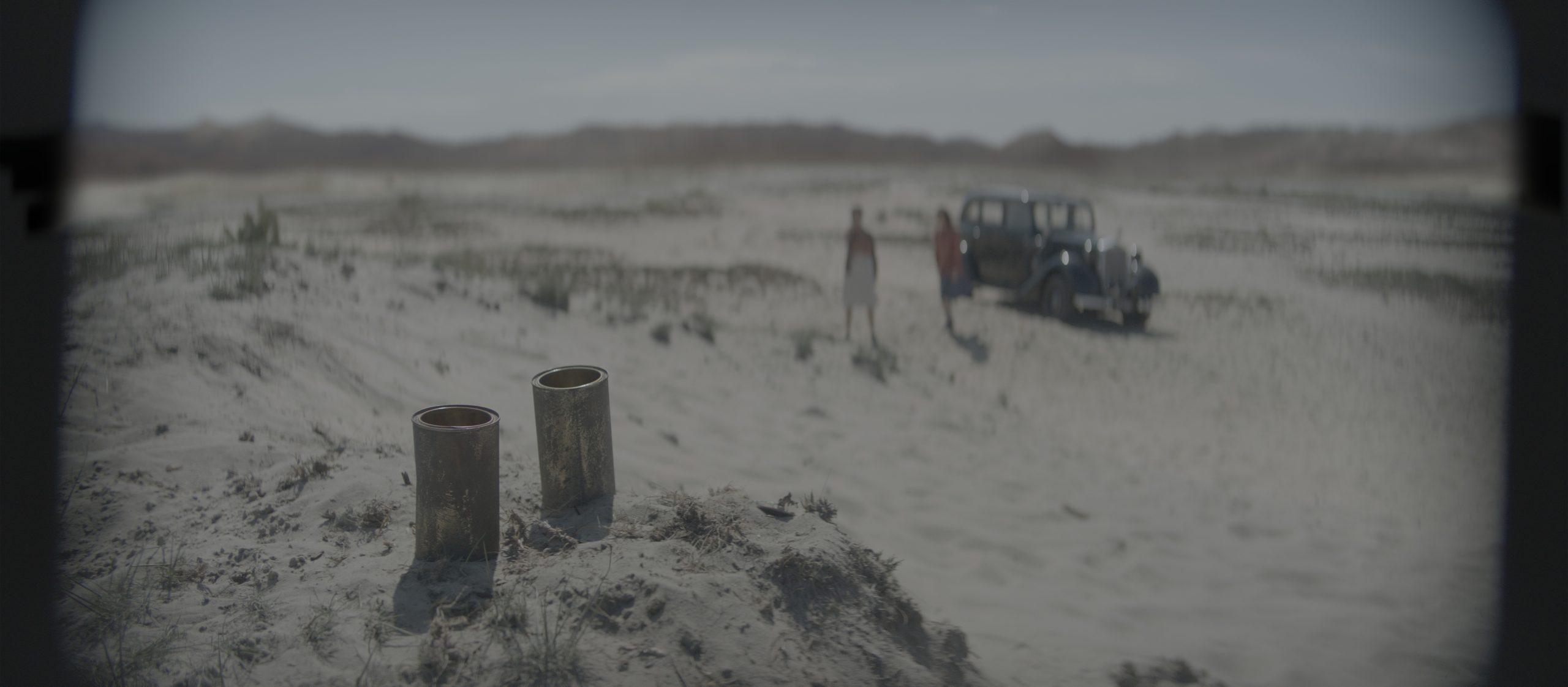 two cylindrical structures stand in a sandy landscape with a blurred figure of a person nearby in the background an old vehicle is parked on the sandy terrain under a clear sky with distant hills digital production Two cylindrical structures stand in a sandy landscape, with a blurred figure of a person nearby. In the background, an old vehicle is parked on the sandy terrain, under a clear sky with distant hills.