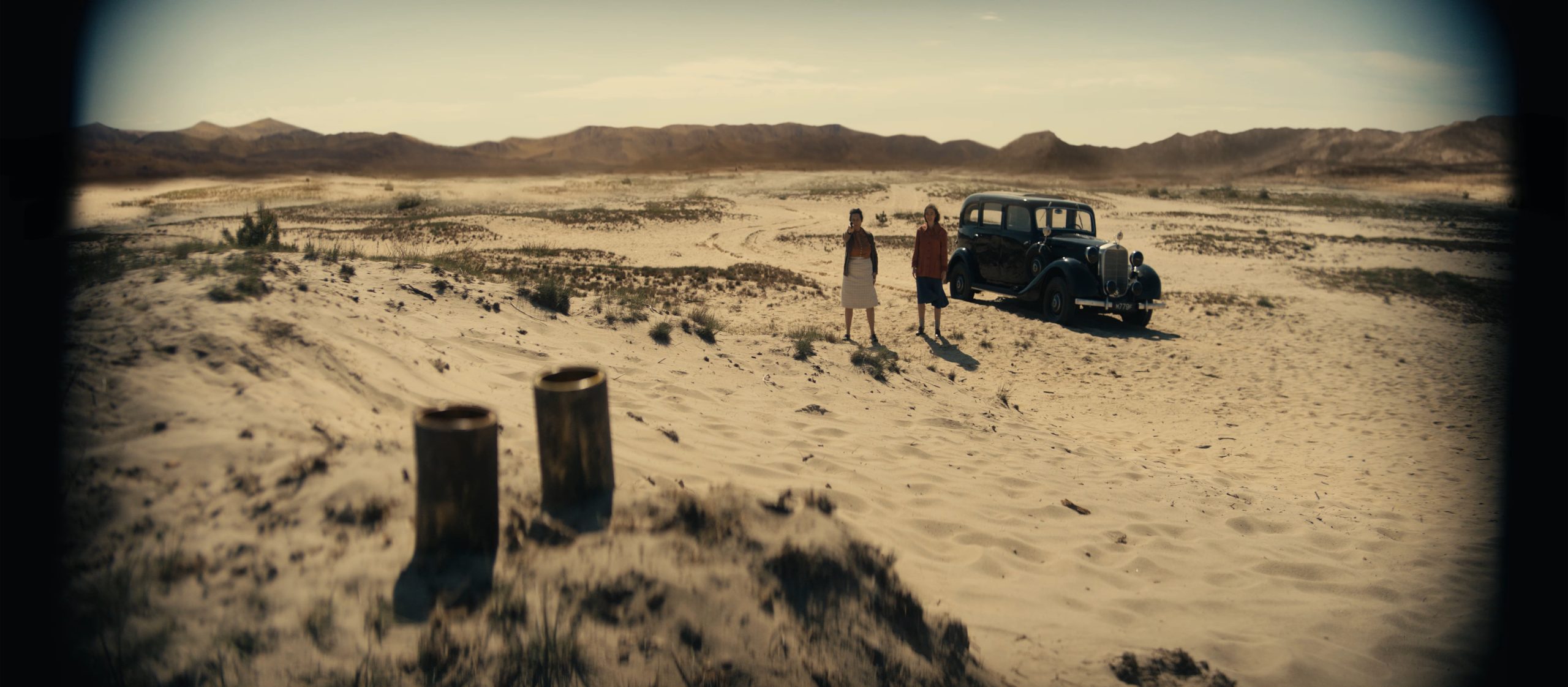 two figures stand in a desolate desert landscape beside an old black car sparse vegetation is visible in the sandy terrain with distant mountains under a clear sky digital production Two figures stand in a desolate desert landscape beside an old black car. Sparse vegetation is visible in the sandy terrain, with distant mountains under a clear sky.