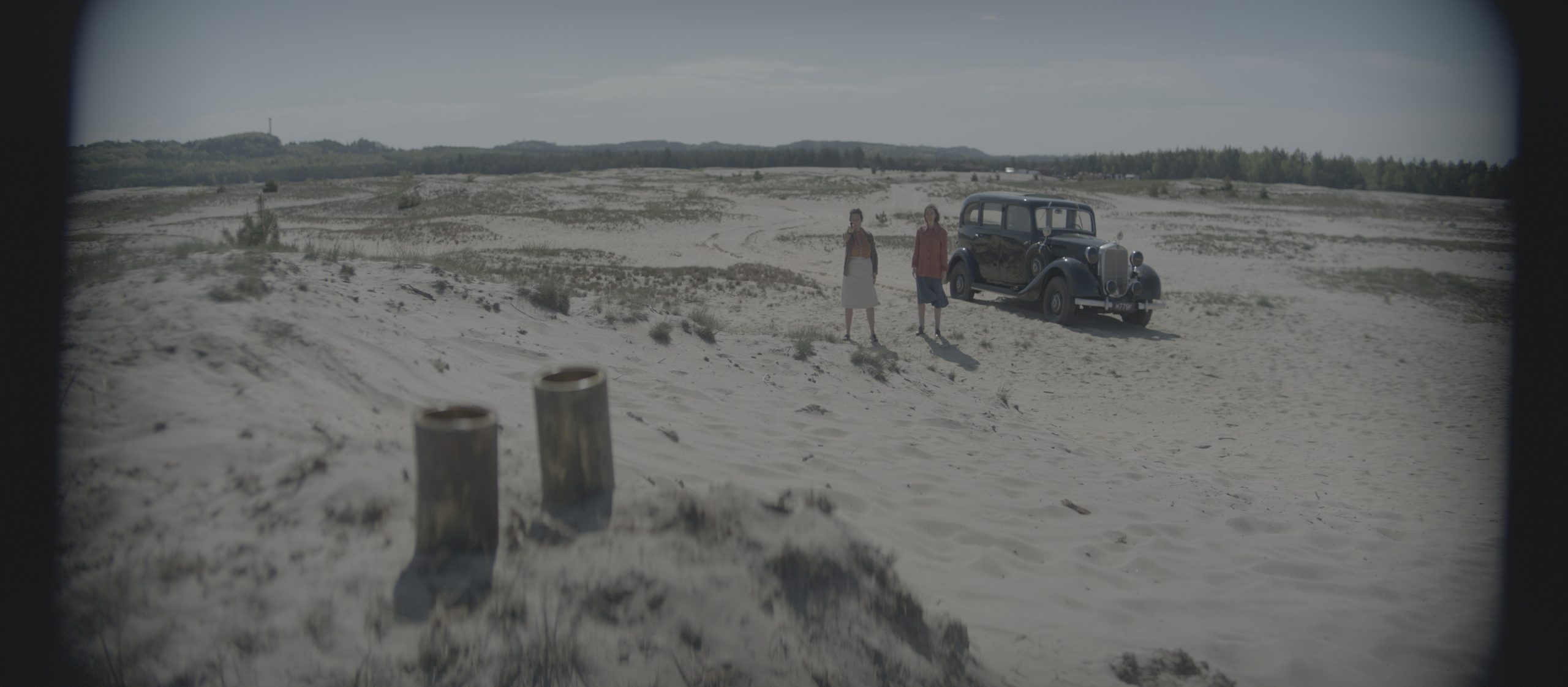 two figures standing on a sandy landscape with sparse vegetation next to a vintage car parked nearby in the foreground there are two cylindrical containers the background features a distant forest and an overcast sky creating a desolate atmosphere digital production Two figures standing on a sandy landscape with sparse vegetation, next to a vintage car parked nearby. In the foreground, there are two cylindrical containers. The background features a distant forest and an overcast sky, creating a desolate atmosphere.