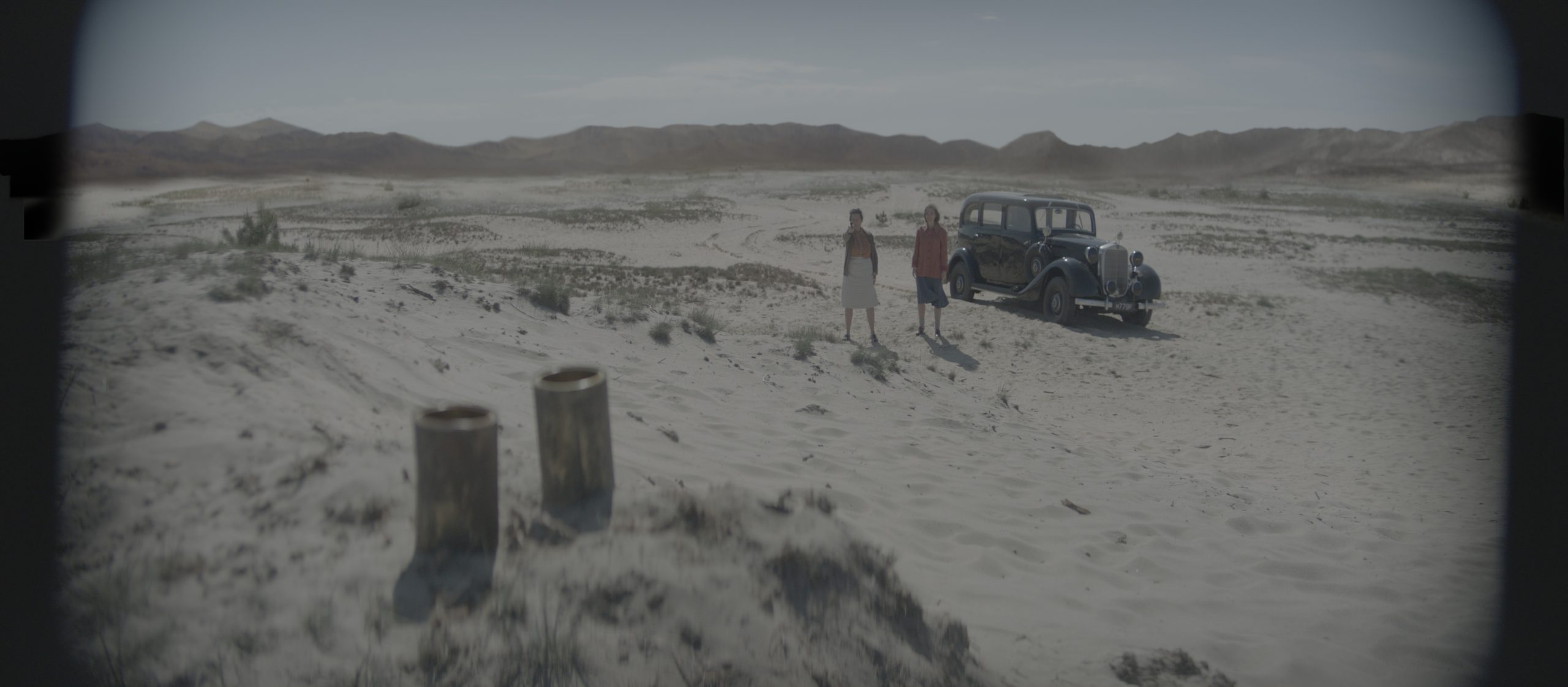 two people standing in a barren landscape with a vintage car nearby the dry sandy terrain is framed by distant hills under a clear sky creating a sense of isolation digital production Two people standing in a barren landscape with a vintage car nearby. The dry, sandy terrain is framed by distant hills under a clear sky, creating a sense of isolation.