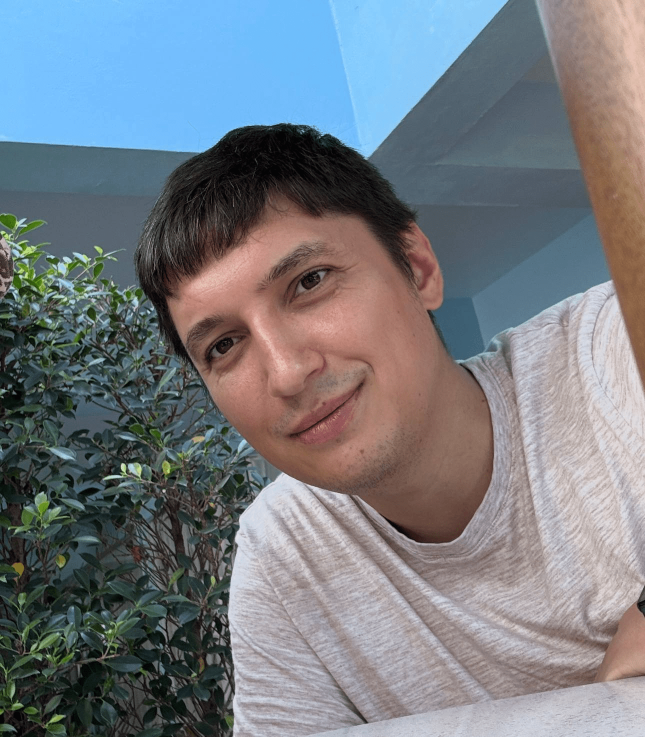 a young man smiling while leaning on a table surrounded by lush green foliage the background features a blue wall and a bright blue sky creating a vibrant cheerful atmosphere digital production A young man smiling while leaning on a table, surrounded by lush green foliage. The background features a blue wall and a bright blue sky, creating a vibrant, cheerful atmosphere.