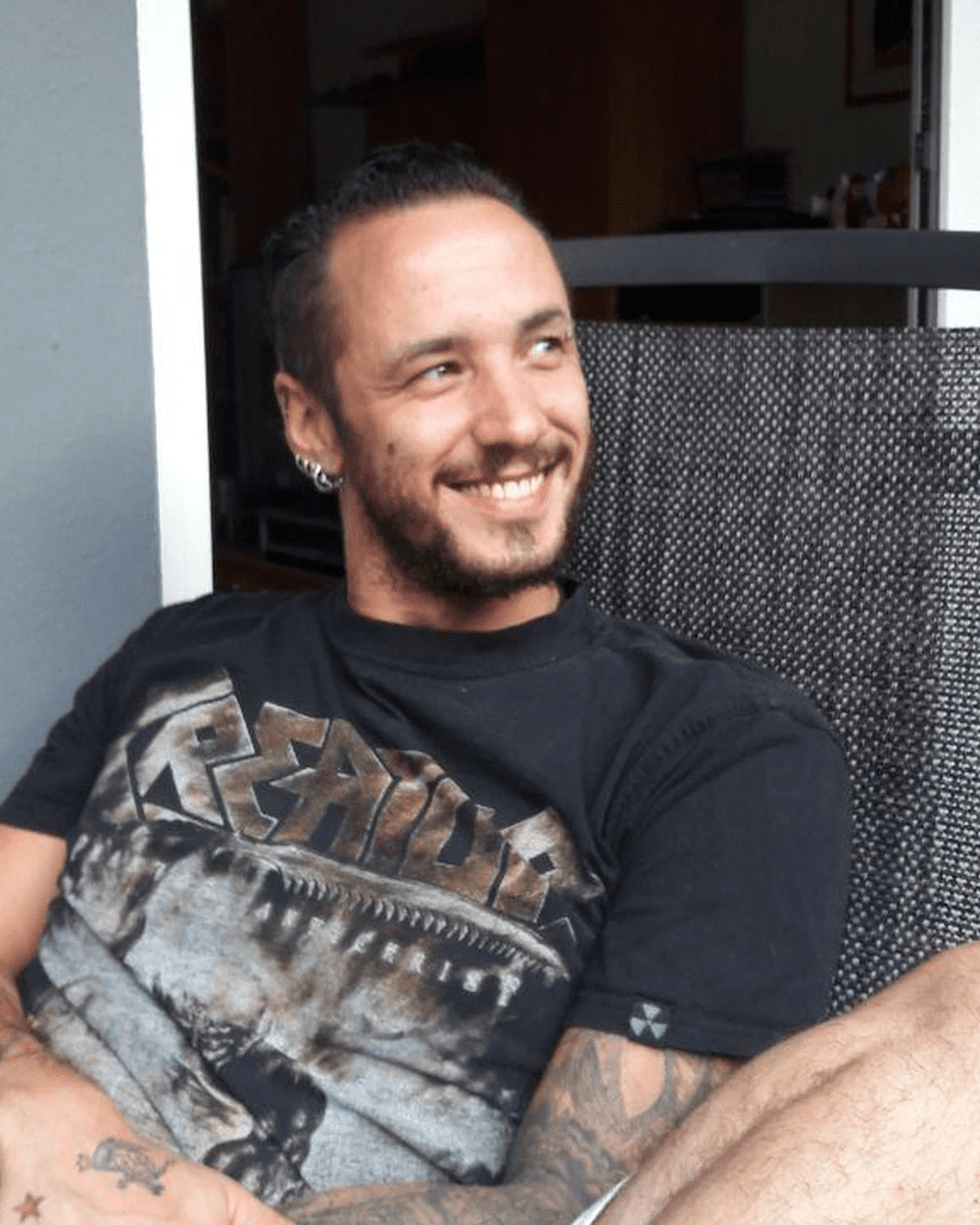 a man with tattoos and earrings smiling while sitting in a chair wearing a black band tshirt the background features a cozy indoor environment with soft lighting digital production A man with tattoos and earrings smiling while sitting in a chair, wearing a black band t-shirt. The background features a cozy indoor environment with soft lighting.