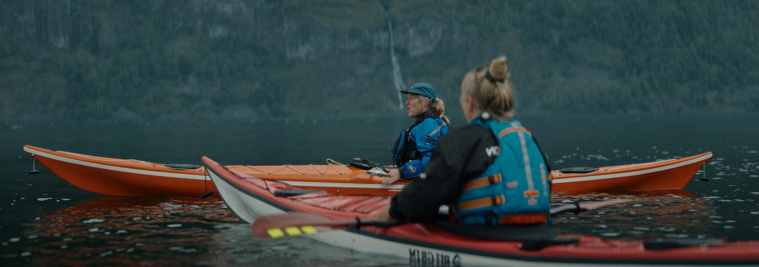 two women kayaking on a calm body of water surrounded by mountains one in a red kayak and the other in an orange kayak they are wearing life jackets and appear engaged in conversation digital production Two women kayaking on a calm body of water surrounded by mountains, one in a red kayak and the other in an orange kayak. They are wearing life jackets and appear engaged in conversation.