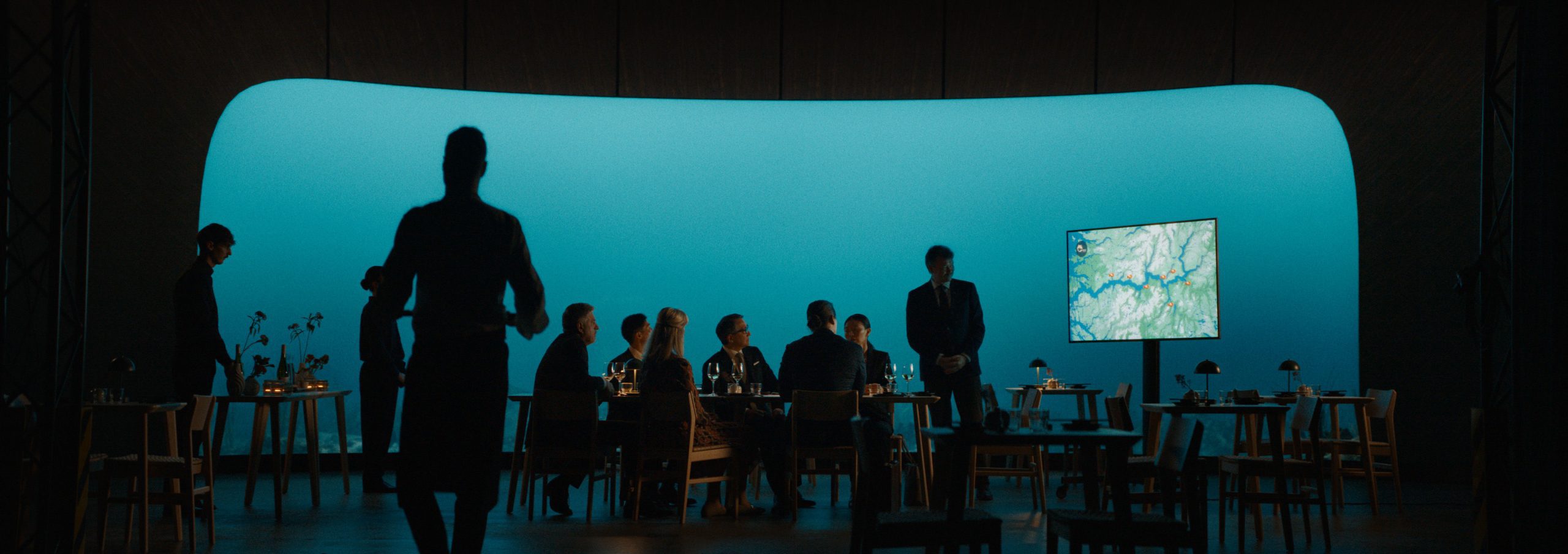 a silhouette of a server stands in the foreground facing a formal dining setup with guests seated around a table a large screen displays a maplike image against a soft blue backdrop creating a modern and elegant atmosphere digital production A silhouette of a server stands in the foreground, facing a formal dining setup with guests seated around a table. A large screen displays a map-like image against a soft blue backdrop, creating a modern and elegant atmosphere.