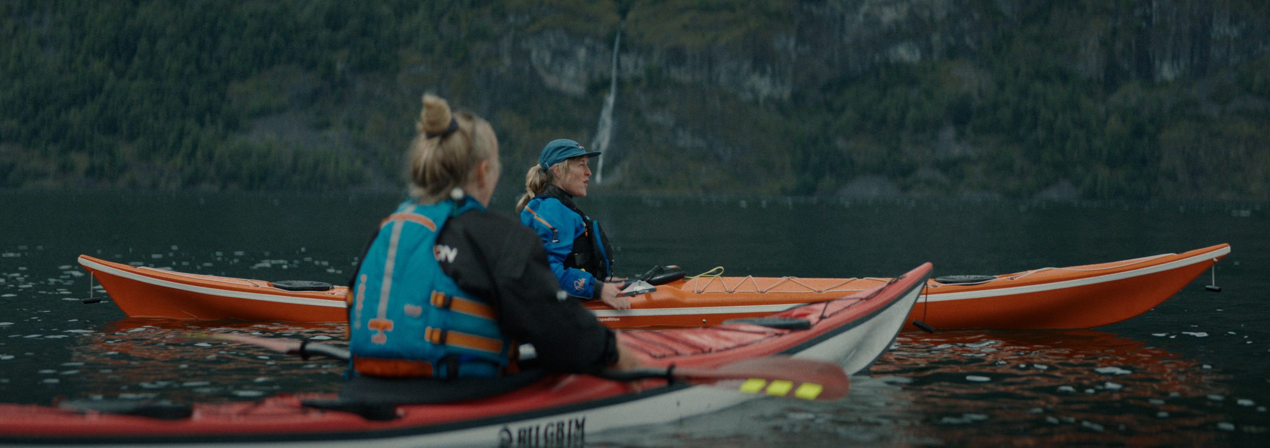 two kayakers on a calm lake one in a red kayak and another in an orange kayak wearing life jackets and paddling in the scenic wilderness with lush green mountains in the background digital production Two kayakers on a calm lake, one in a red kayak and another in an orange kayak, wearing life jackets and paddling in the scenic wilderness, with lush green mountains in the background.