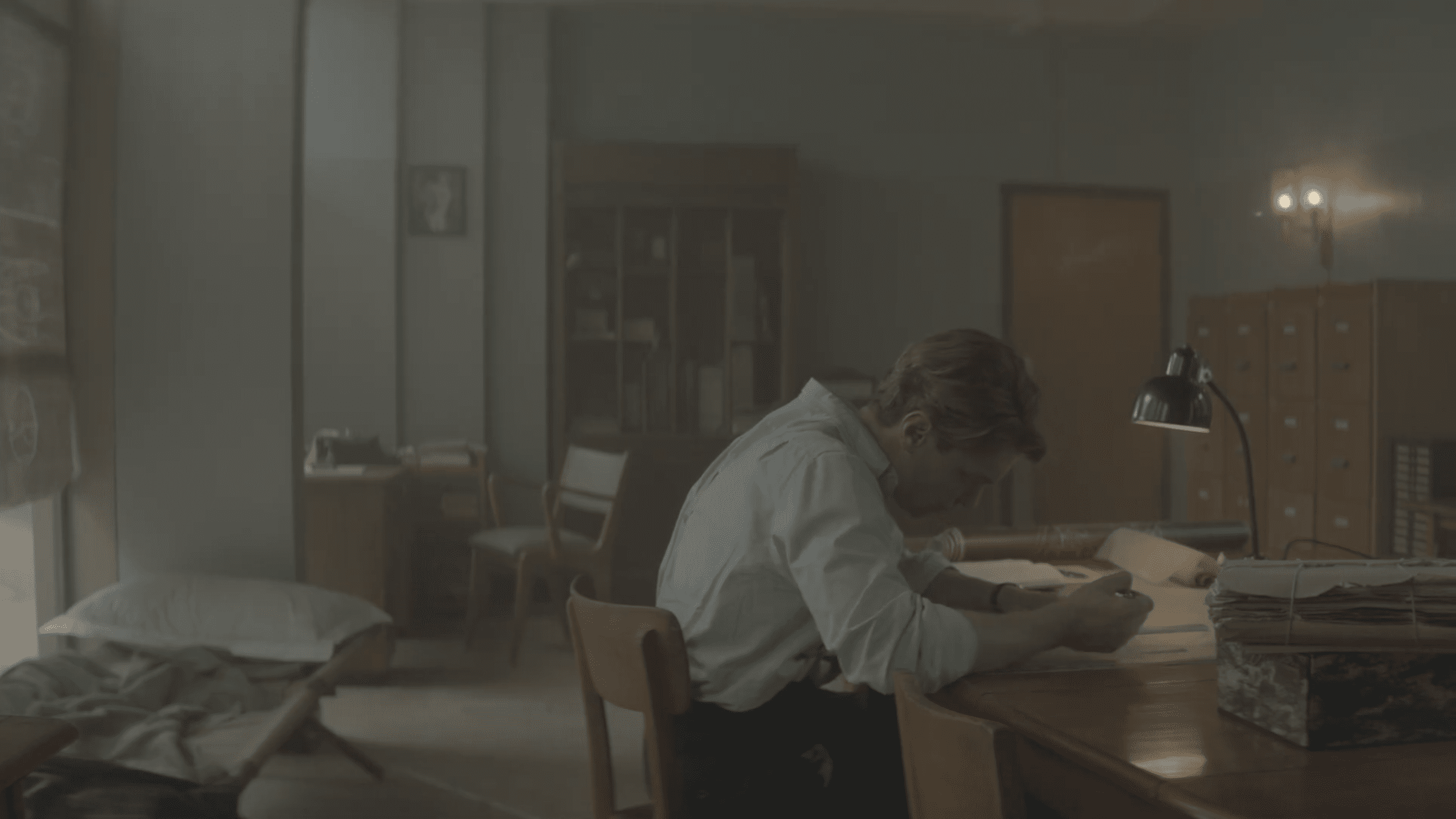 a young man in a white shirt sits pensively at a desk surrounded by papers in a dimly lit room with muted colors a wooden cabinet and a cozy chair are visible in the background conveying a reflective atmosphere digital production A young man in a white shirt sits pensively at a desk, surrounded by papers, in a dimly lit room with muted colors. A wooden cabinet and a cozy chair are visible in the background, conveying a reflective atmosphere.