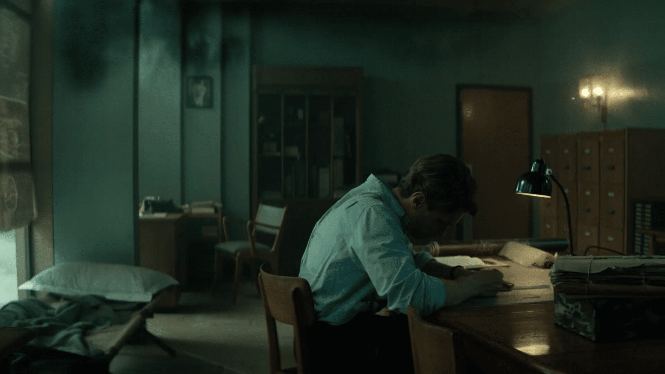a man in a light blue shirt sits at a wooden desk focused on writing the room is dimly lit with shadows on the walls and a messy appearance a bed and filing cabinets can be seen in the background creating an atmosphere of solitude digital production A man in a light blue shirt sits at a wooden desk, focused on writing. The room is dimly lit, with shadows on the walls and a messy appearance. A bed and filing cabinets can be seen in the background, creating an atmosphere of solitude.