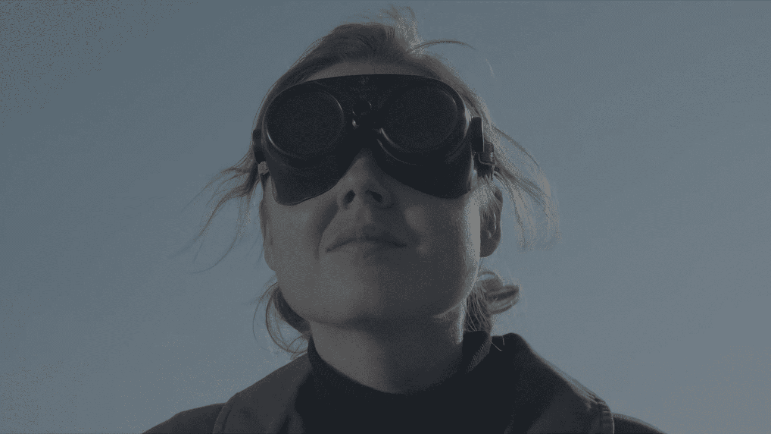 a woman wearing dark goggles gazes upwards against a pale sky with her hair slightly tousled by the wind she is dressed in a dark outfit conveying a sense of determination and focus digital production A woman wearing dark goggles gazes upwards against a pale sky, with her hair slightly tousled by the wind. She is dressed in a dark outfit, conveying a sense of determination and focus.