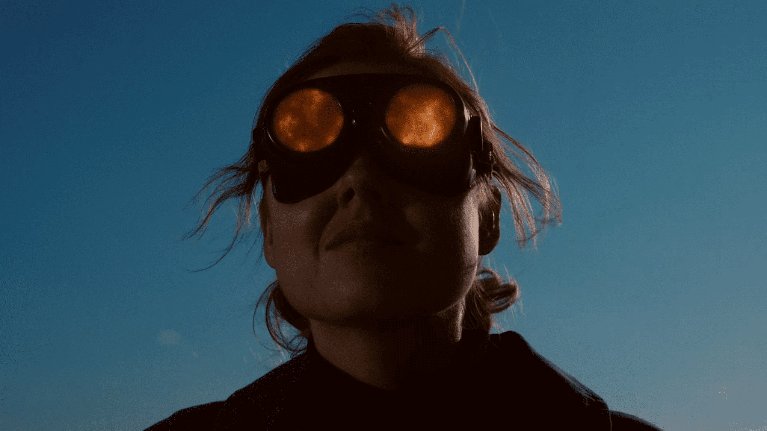 a person wearing large reflective goggles that show a fiery orange sky with their hair gently blowing in the wind against a blue background digital production A person wearing large, reflective goggles that show a fiery orange sky, with their hair gently blowing in the wind against a blue background.