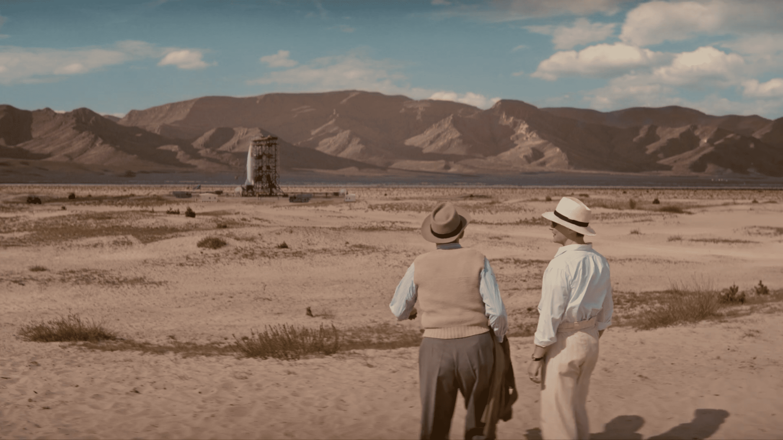 two men in vintage attire stand on arid land gazing at a weathered wooden structure in the distance the backdrop features rugged mountains under a blue sky with scattered clouds digital production Two men in vintage attire stand on arid land, gazing at a weathered wooden structure in the distance. The backdrop features rugged mountains under a blue sky with scattered clouds.