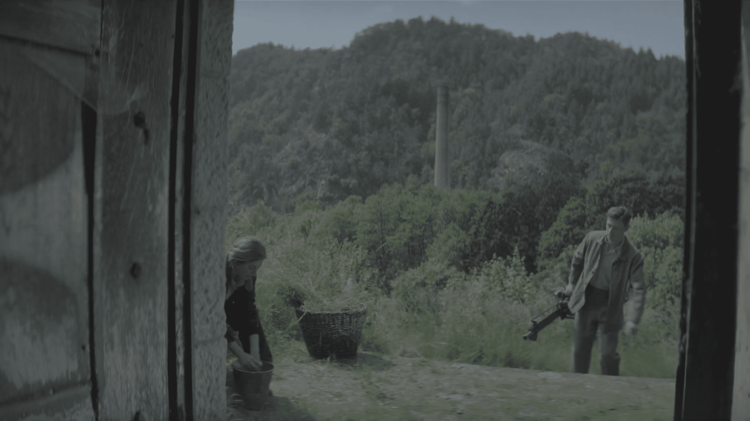 a dimly lit scene viewed through an open door showing a woman crouched by a basket and a man standing nearby with a weapon surrounded by lush green hills under a cloudy sky digital production A dimly lit scene viewed through an open door, showing a woman crouched by a basket and a man standing nearby with a weapon, surrounded by lush green hills under a cloudy sky.