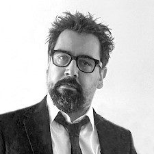 a man with a beard and glasses wearing a dark suit jacket over a white shirt poses for a portrait against a plain background conveying a professional look digital production A man with a beard and glasses, wearing a dark suit jacket over a white shirt, poses for a portrait against a plain background, conveying a professional look.