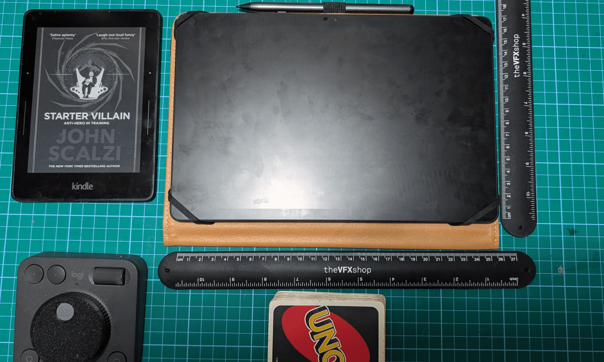 a flat lay image featuring a kindle displaying the book starter villain by john scalzi a tablet on a leather surface a round remote a game card and various rulers all arranged on a green cutting mat digital production A flat lay image featuring a Kindle displaying the book 'Starter Villain' by John Scalzi, a tablet on a leather surface, a round remote, a game card, and various rulers, all arranged on a green cutting mat.