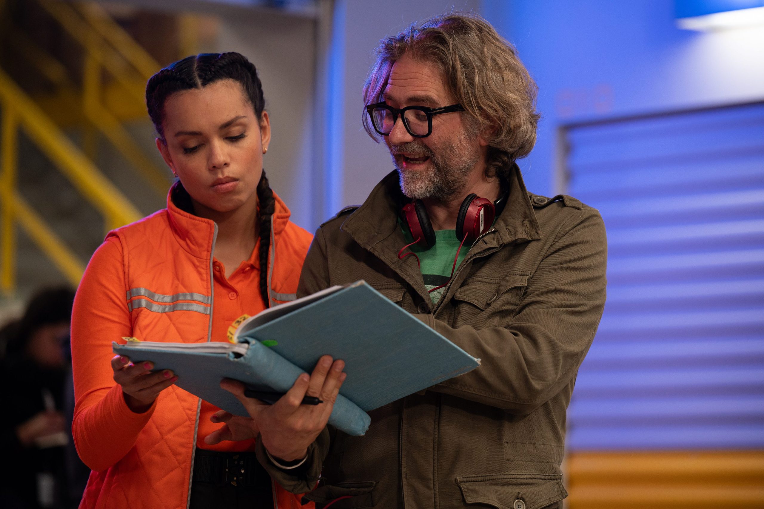 a director discussing a script with a young female actor on a set the actor wears an orange jacket and the director in a green shirt and beige jacket holds a blue folder the background shows a welllit film set digital production A director discussing a script with a young female actor on a set. The actor wears an orange jacket, and the director, in a green shirt and beige jacket, holds a blue folder. The background shows a well-lit film set.