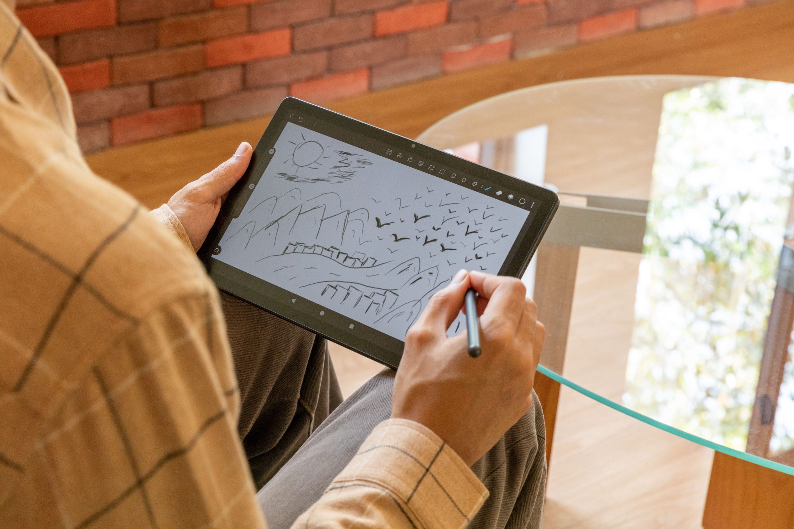 a person sitting at a glass table drawing on a digital tablet the screen displays a pen sketch of a landscape with mountains a sun and birds while the brick wall in the background adds warmth to the scene digital production A person sitting at a glass table, drawing on a digital tablet. The screen displays a pen sketch of a landscape with mountains, a sun, and birds, while the brick wall in the background adds warmth to the scene.