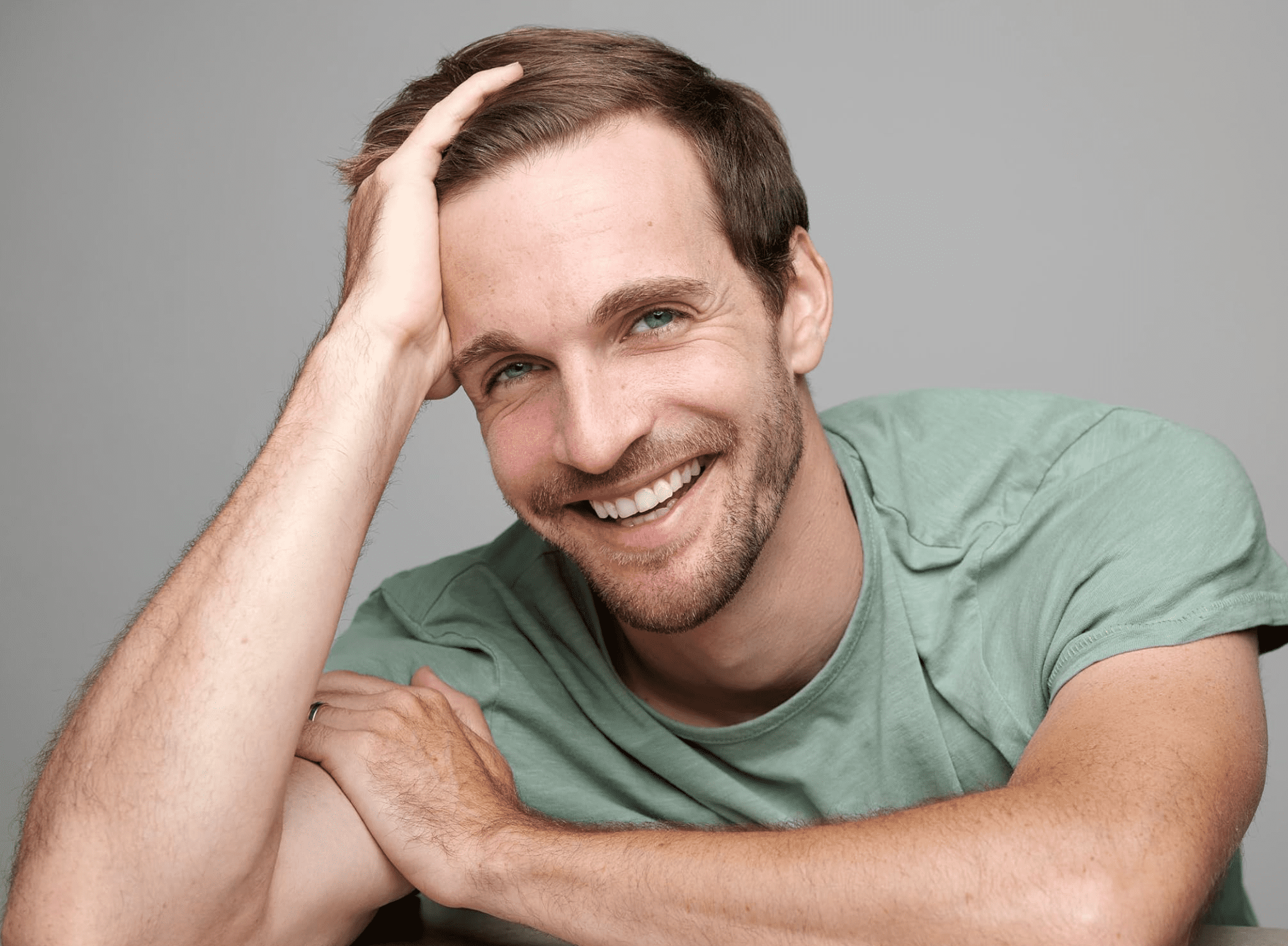 a smiling man with light brown hair and blue eyes rests his hand on his head while leaning on a table he wears a casual green tshirt and the background is a soft gray creating a warm friendly atmosphere digital production A smiling man with light brown hair and blue eyes rests his hand on his head while leaning on a table. He wears a casual green T-shirt, and the background is a soft gray, creating a warm, friendly atmosphere.
