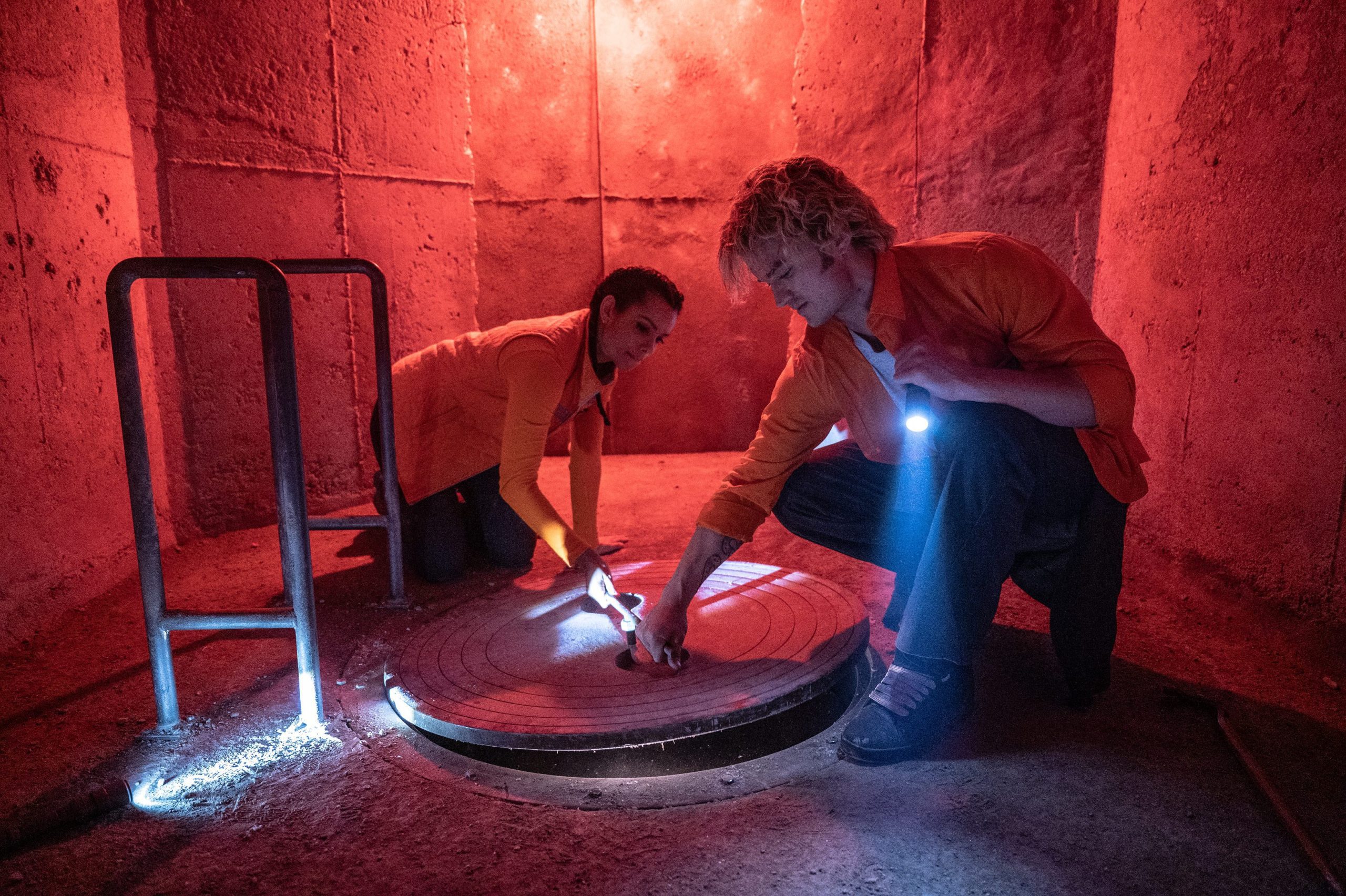 two individuals in matching orange shirts are kneeling on the floor of a dimly lit redtinted environment one person is using a flashlight to illuminate a circular object on the ground while the other is focused on the task at hand digital production Two individuals in matching orange shirts are kneeling on the floor of a dimly lit, red-tinted environment. One person is using a flashlight to illuminate a circular object on the ground, while the other is focused on the task at hand.