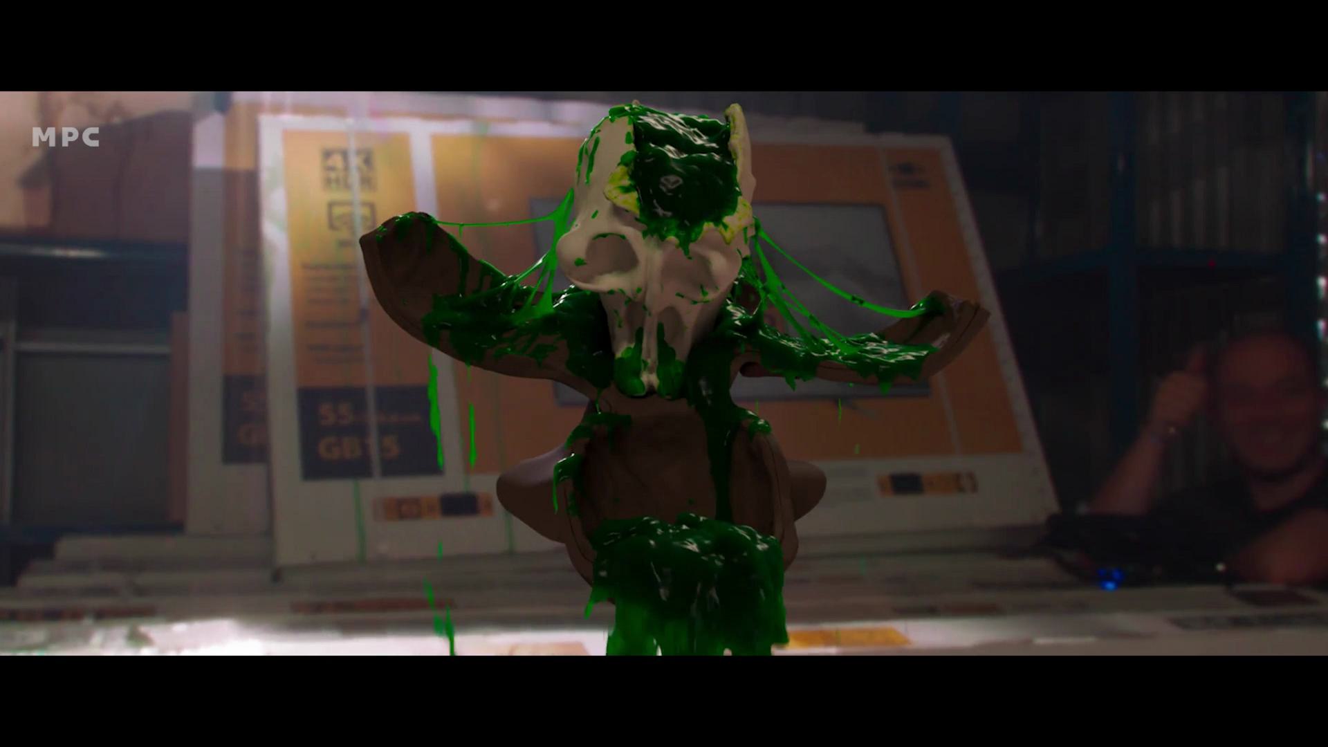 a small statue with a skull design is covered in bright green slime sitting on a cluttered table in the background blurred shapes suggest the presence of people and equipment digital production A small statue with a skull design is covered in bright green slime, sitting on a cluttered table. In the background, blurred shapes suggest the presence of people and equipment.