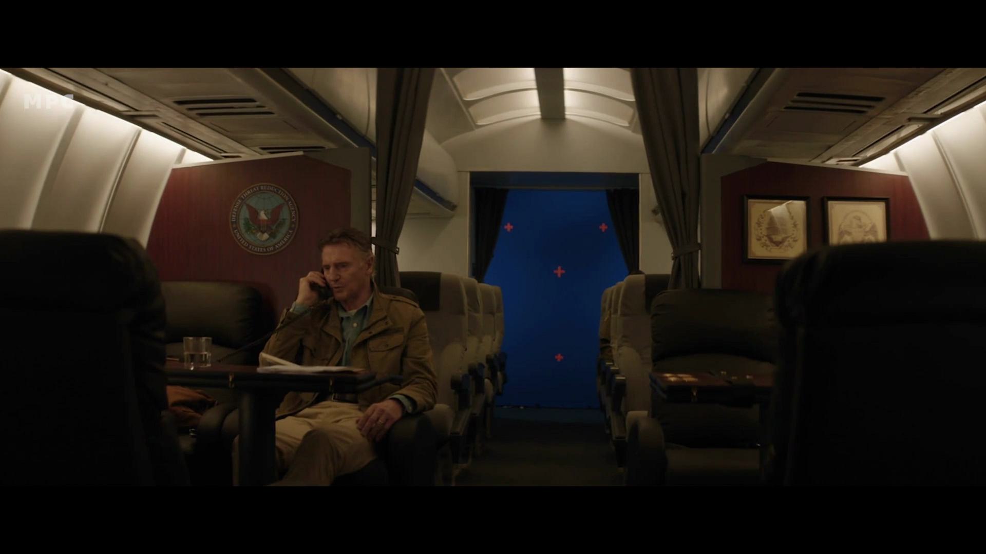 an empty airplane cabin with dim lighting a man in a military jacket sits at a table talking on the phone the walls display the military emblem and there are two framed pictures the rear exit has a blue backdrop visible through the door digital production An empty airplane cabin with dim lighting. A man in a military jacket sits at a table, talking on the phone. The walls display the military emblem, and there are two framed pictures. The rear exit has a blue backdrop visible through the door.