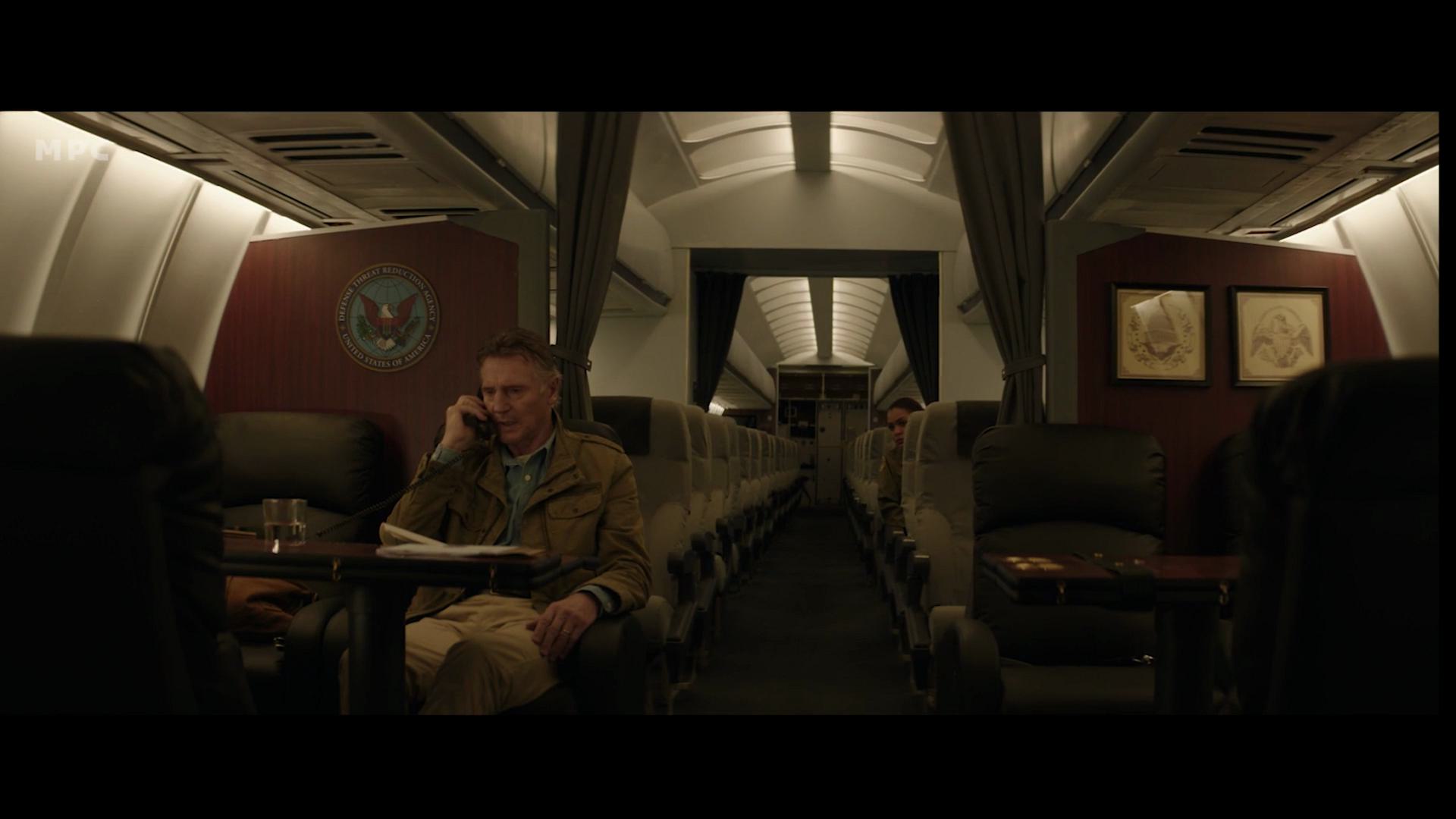a passenger sitting in an airline seat on a dimly lit plane talking on the phone the cabin features plush seats an emblem on the wall and framed artwork with a few empty rows visible in the background digital production A passenger sitting in an airline seat on a dimly lit plane, talking on the phone. The cabin features plush seats, an emblem on the wall, and framed artwork, with a few empty rows visible in the background.