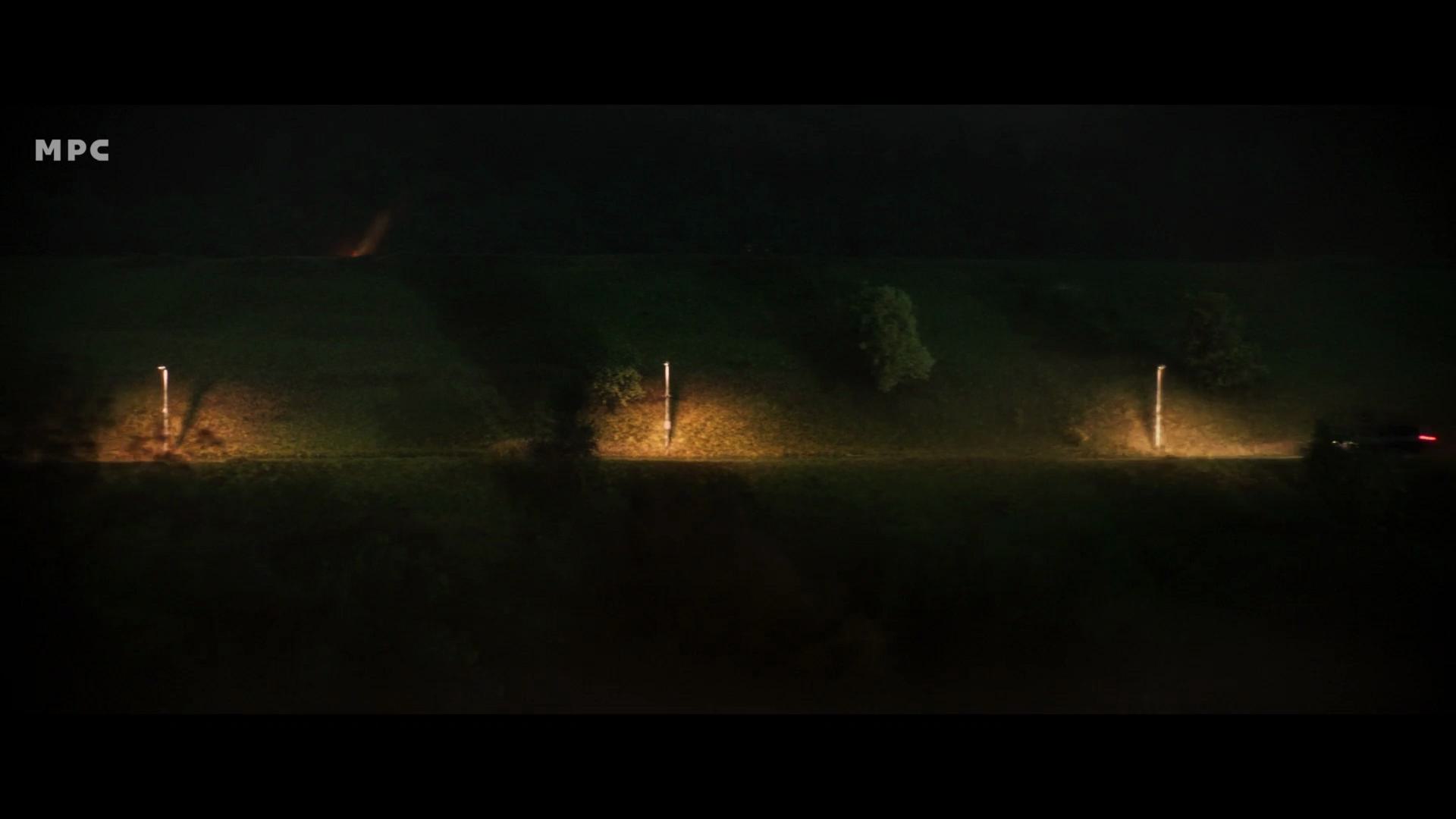 a dimly lit open field at night featuring a meandering road with illuminated posts lining either side a vehicles headlights illuminate the scene casting shadows on nearby trees the atmosphere is serene yet slightly mysterious digital production A dimly lit open field at night, featuring a meandering road with illuminated posts lining either side. A vehicle's headlights illuminate the scene, casting shadows on nearby trees. The atmosphere is serene yet slightly mysterious.